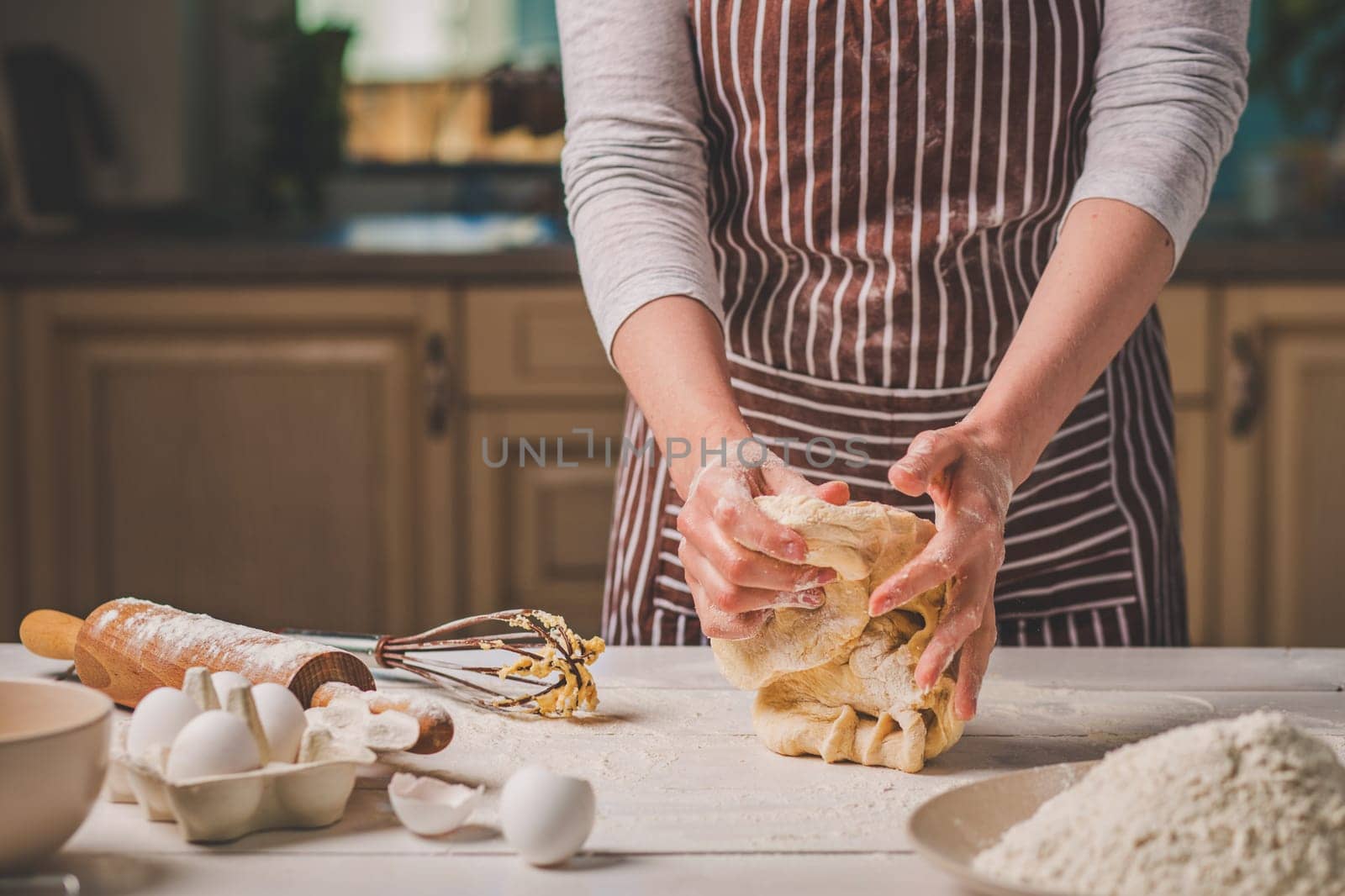 Woman hands kneading dough on kitchen table. A woman in a striped apron is cooking in the kitchen