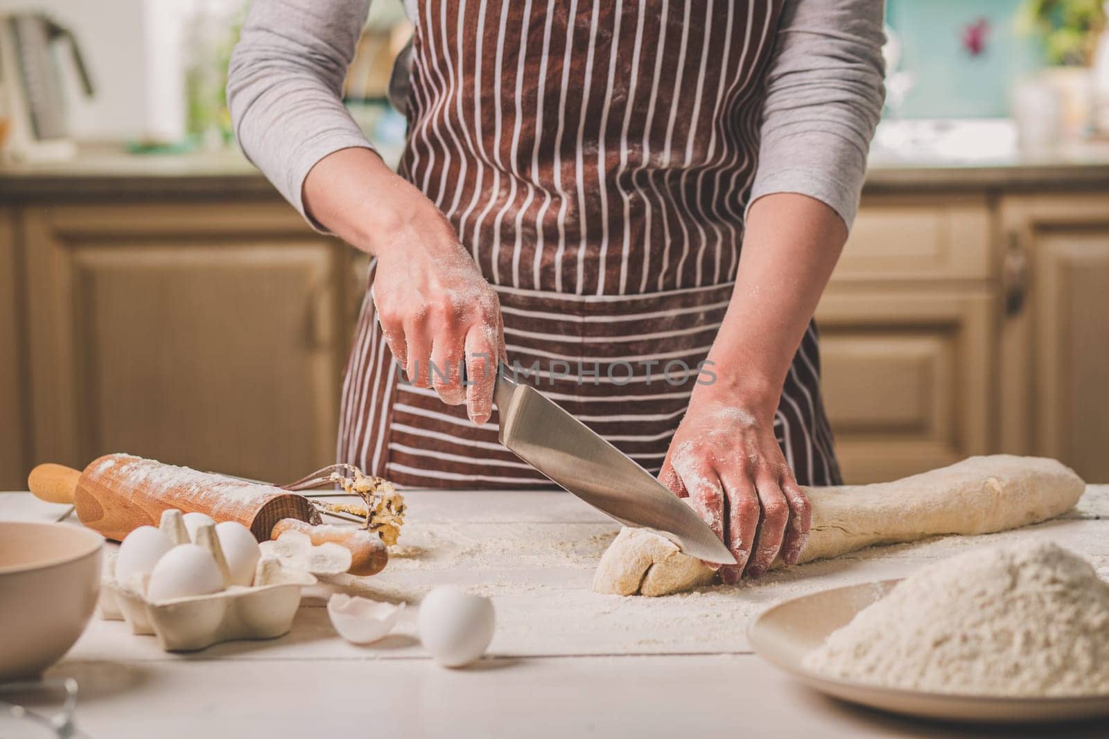 Close-up view of two woman's hands cut knife dough. A woman in a striped apron is cooking in the kitchen