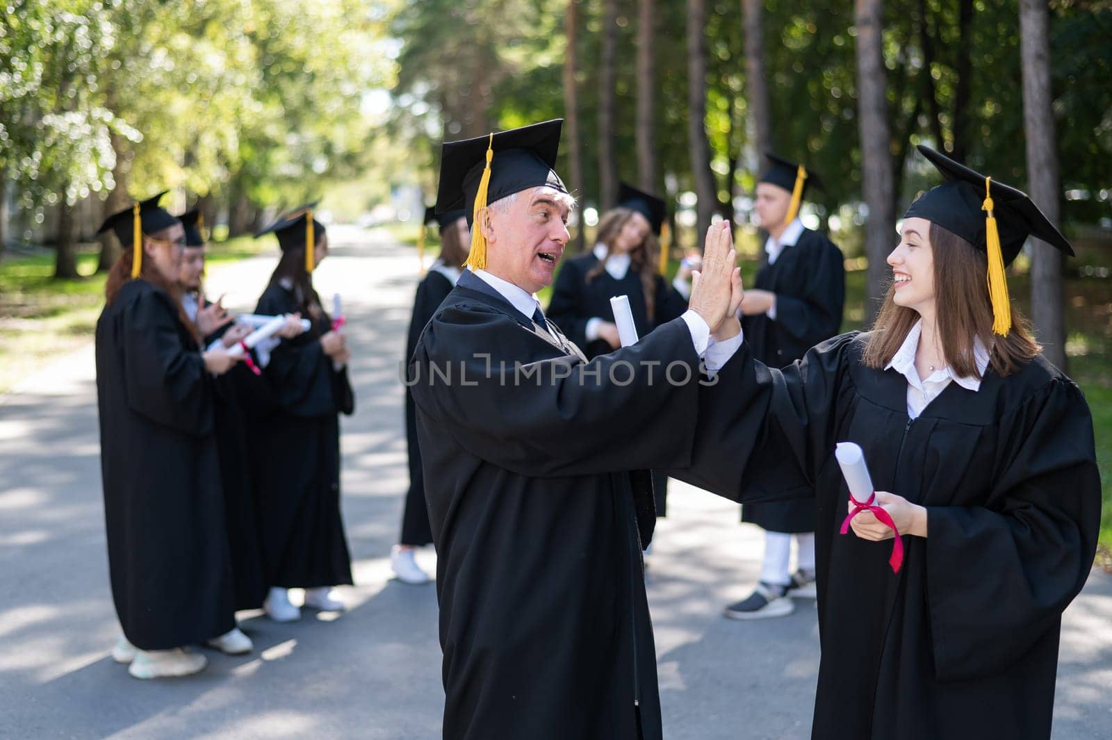 A group of graduates in robes outdoors. An elderly man and a young woman congratulate each other on their graduation