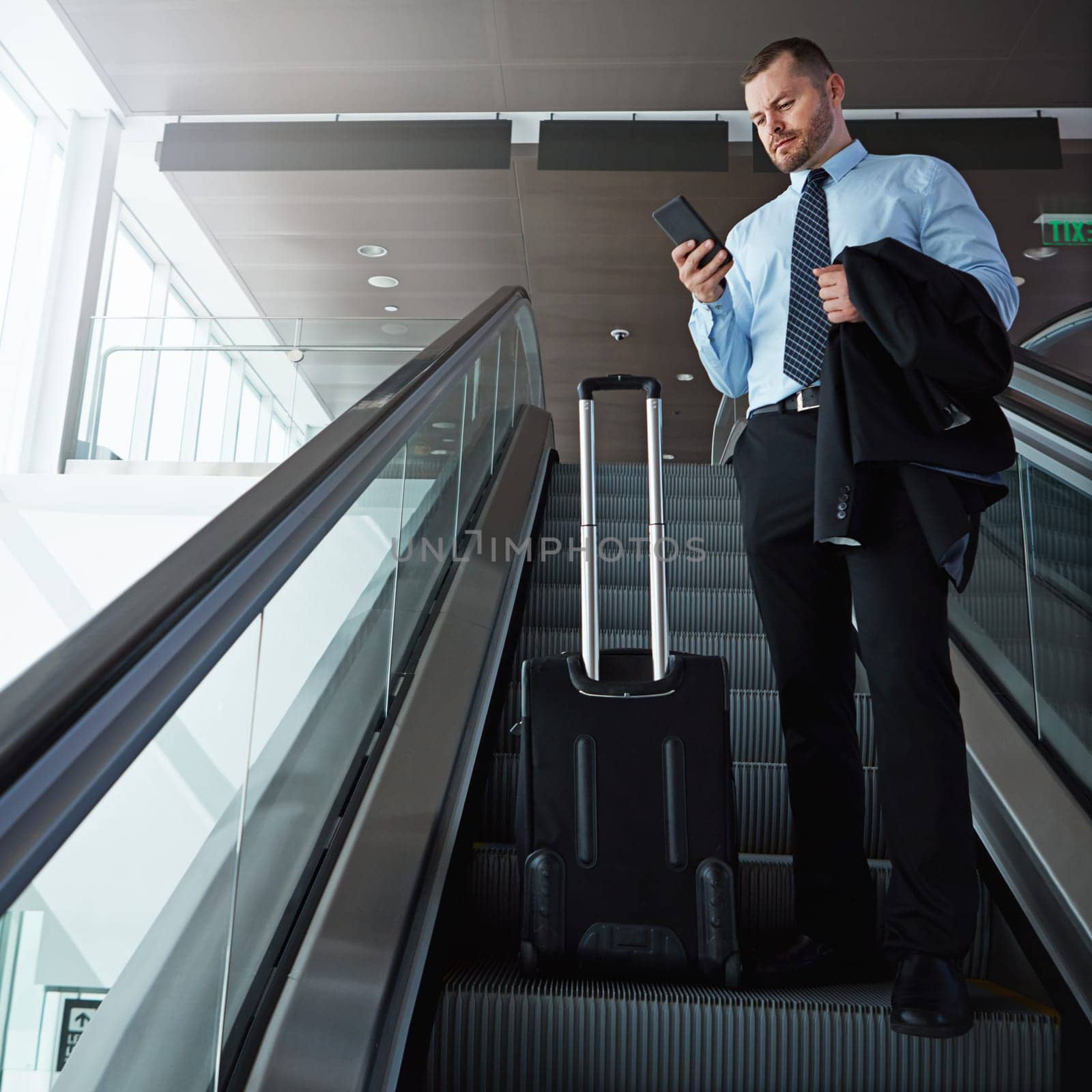 Modern business travel demands modern technology. a businessman using a mobile phone while traveling down an escalator in an airport