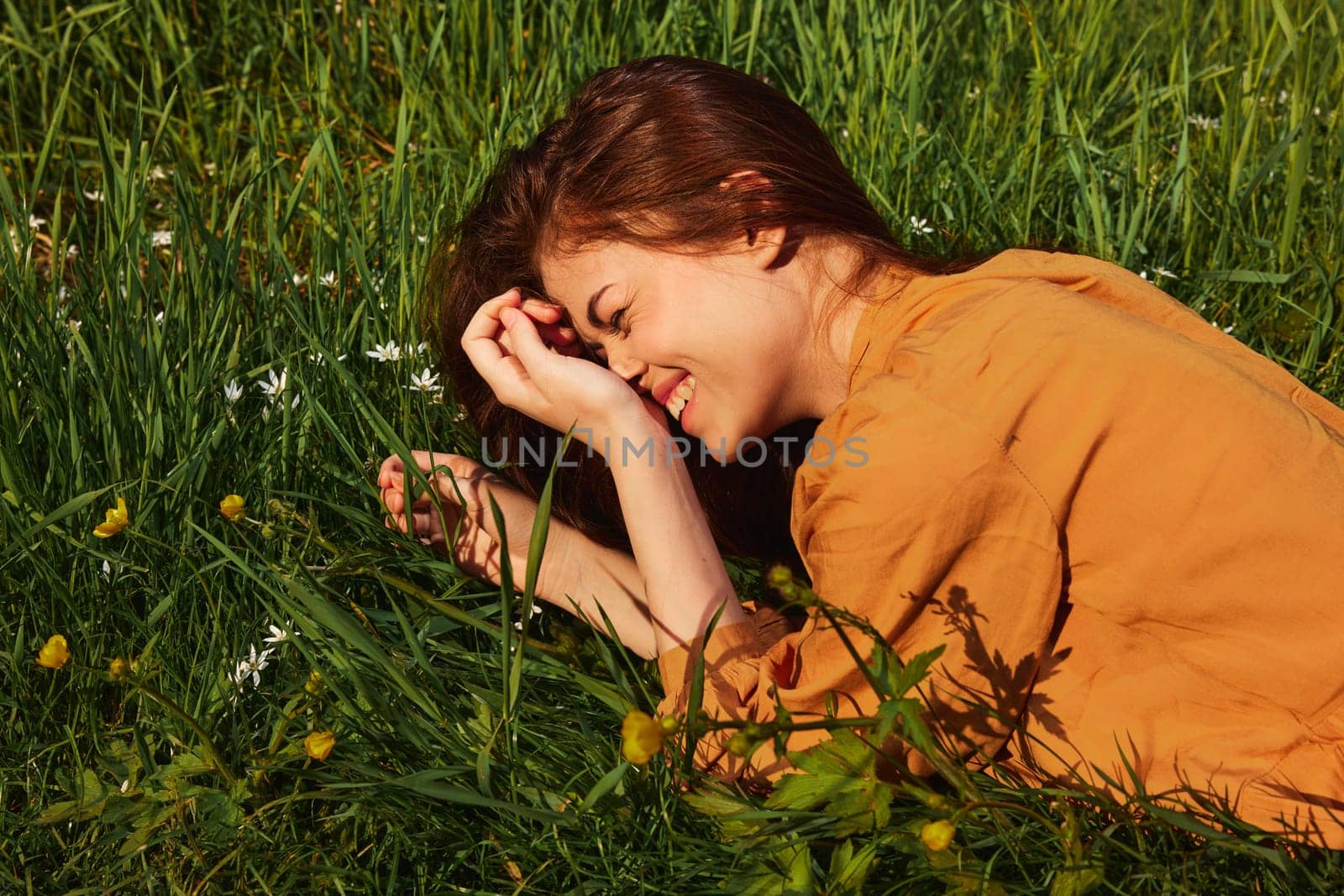 a calm woman with long red hair lies in a green field with yellow flowers, in an orange dress smiling pleasantly, closing her eyes from the bright summer sun. High quality photo
