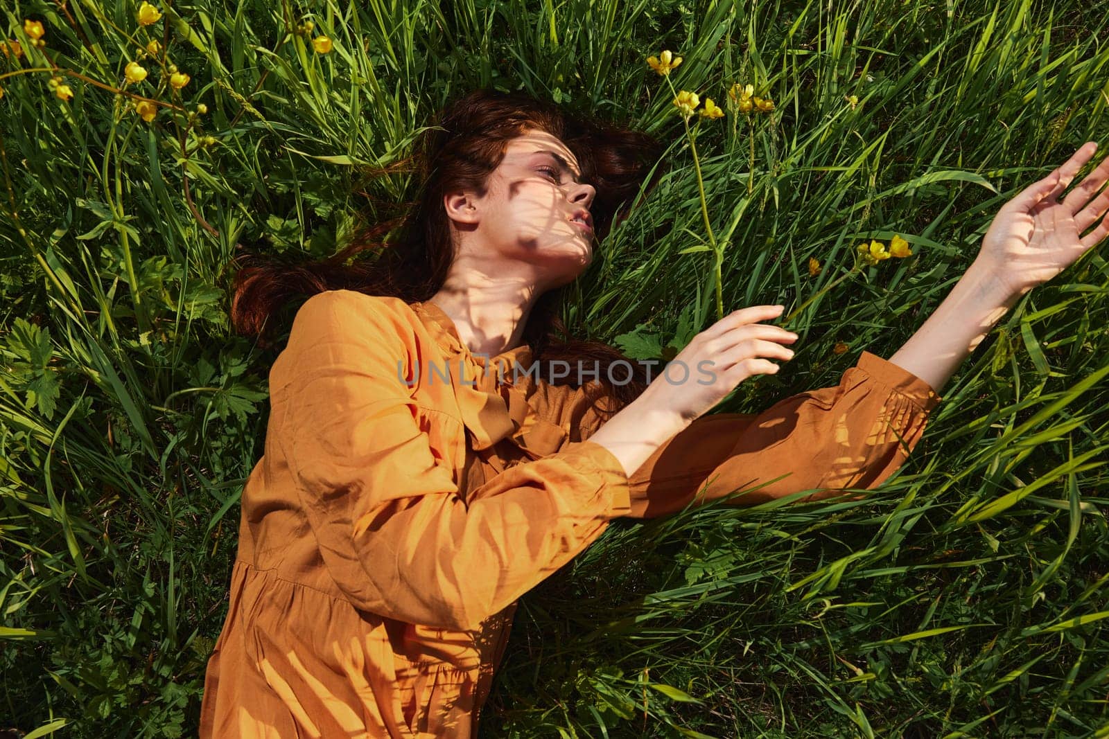 a calm woman with long red hair lies in a green field of tall grass in an orange dress with her eyes closed, stretching her arms in front of her. High quality photo