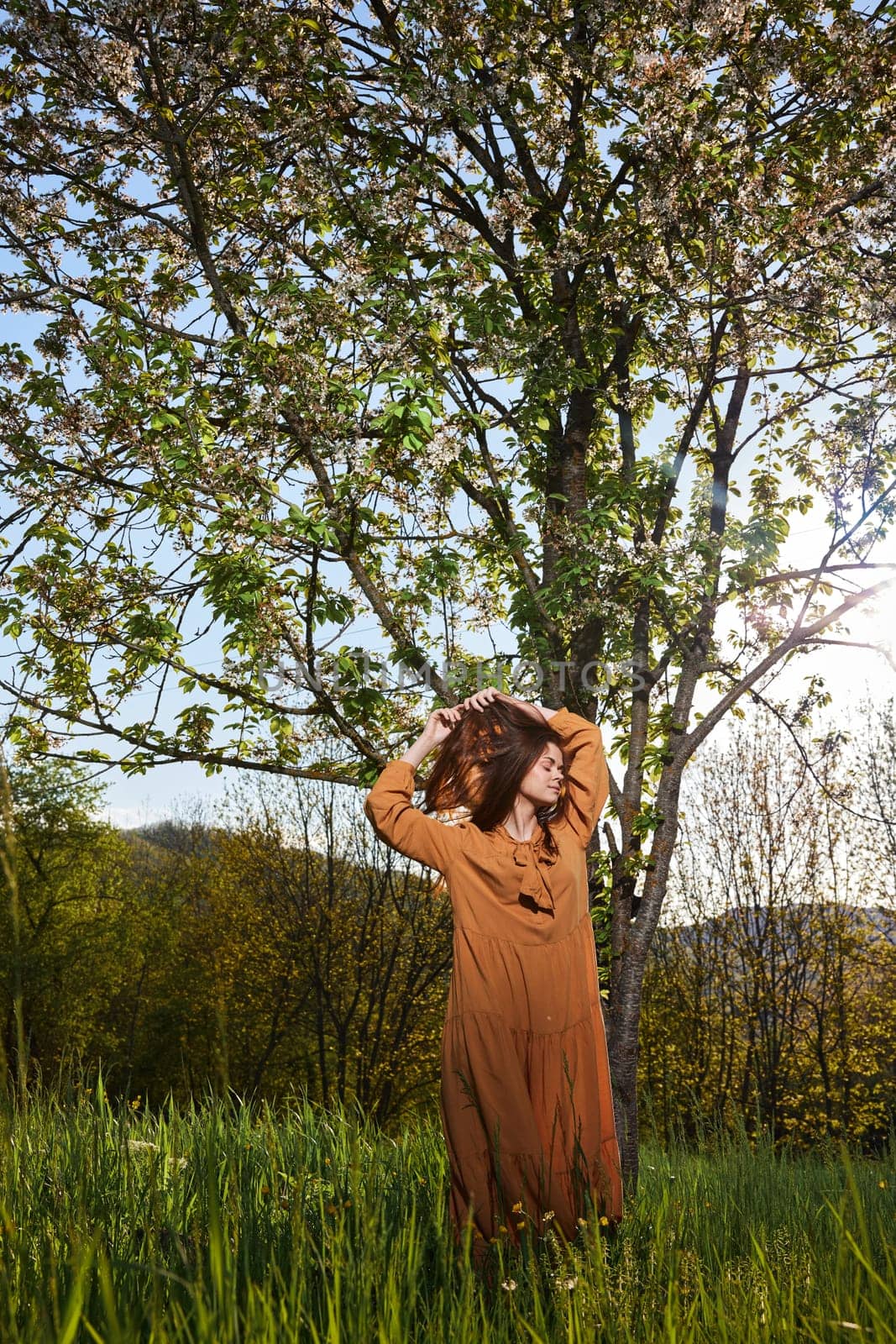 woman with long red hair stands in the countryside near a flowering tree in a long orange dress and holds her hair with her hands while standing sideways to the camera. Vertical photography by Vichizh