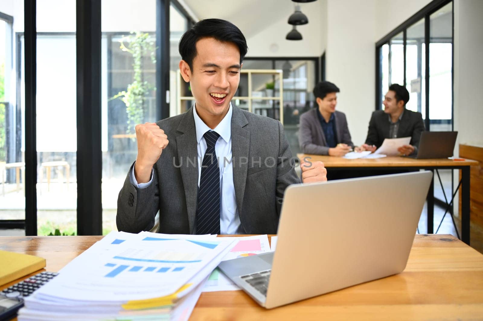 Happy male marketing business manager looking at laptop screen, making winner yes gesture, celebrating business success by prathanchorruangsak