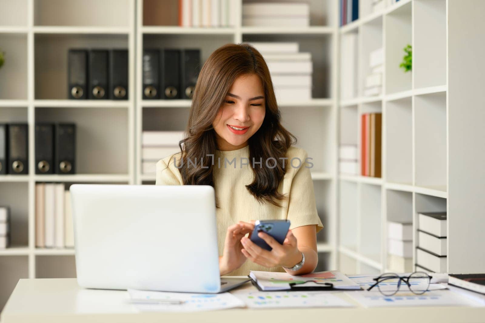 Satisfied asian female financial advisor chatting with client online on smart phone and using laptop at workstation.