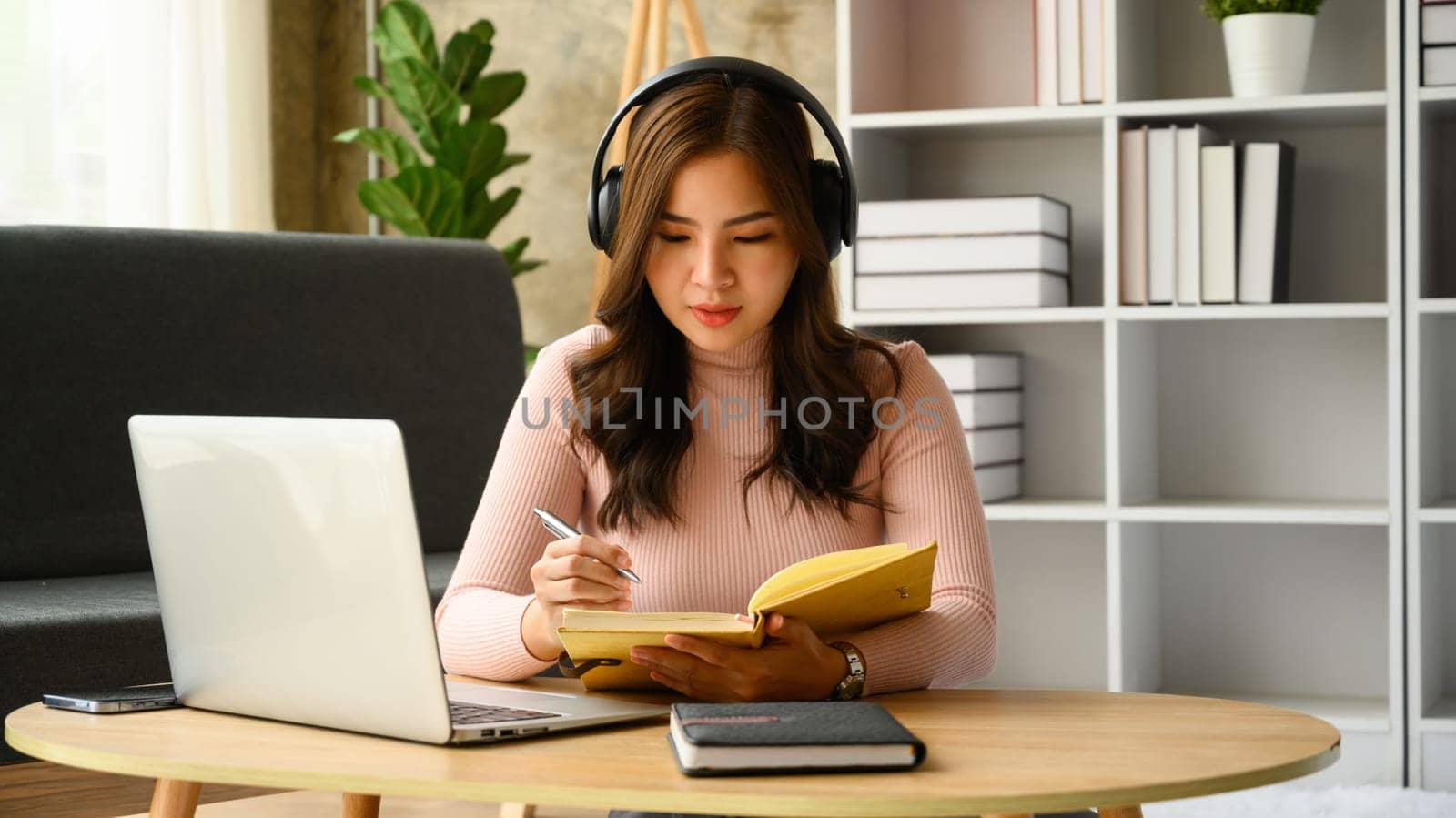 Focused young female student wearing headphones listening online lecture and making notes on notebook.