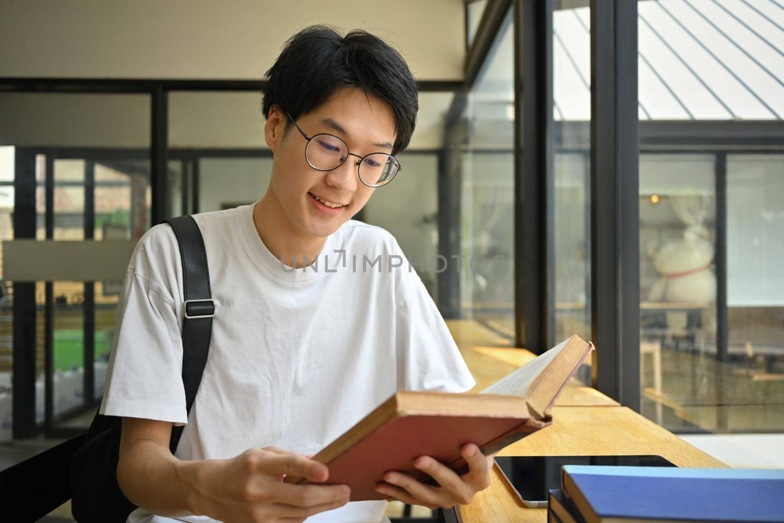 Smiling asian man student in glasses sitting at coffee shop and reading book. Education, learning and technology concept.