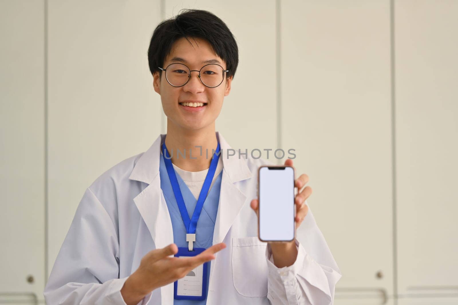 Smiling male doctor in uniform holding smartphone screen, presenting blank smartphone display for medical advertisement, mock up.