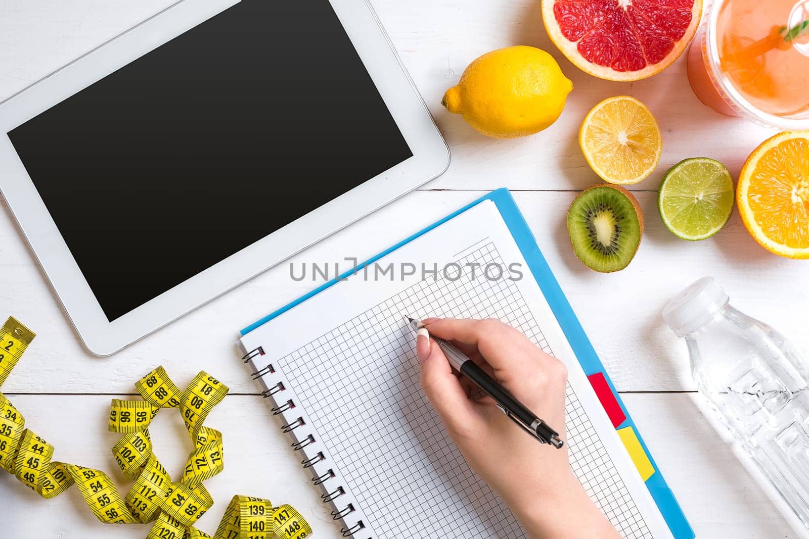 Fresh juice in glass from citrus fruits - lemon, grapefruit, orange, notebook with pencil on white wooden background, top view. White tablet with copy space