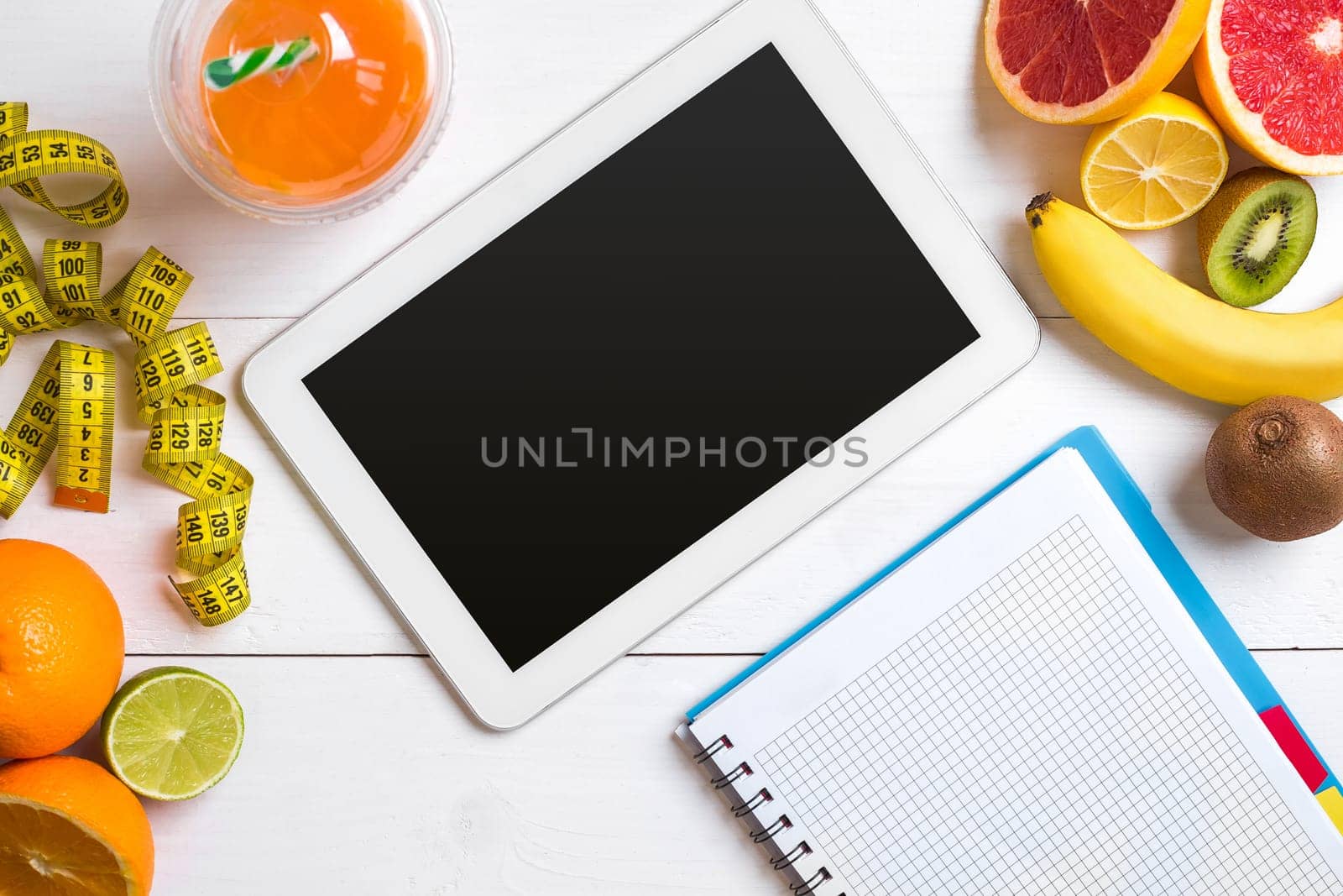 Fresh juice in glass from citrus fruits - lemon, grapefruit, orange, notebook with pencil on white wooden background, top view. White tablet with copy space