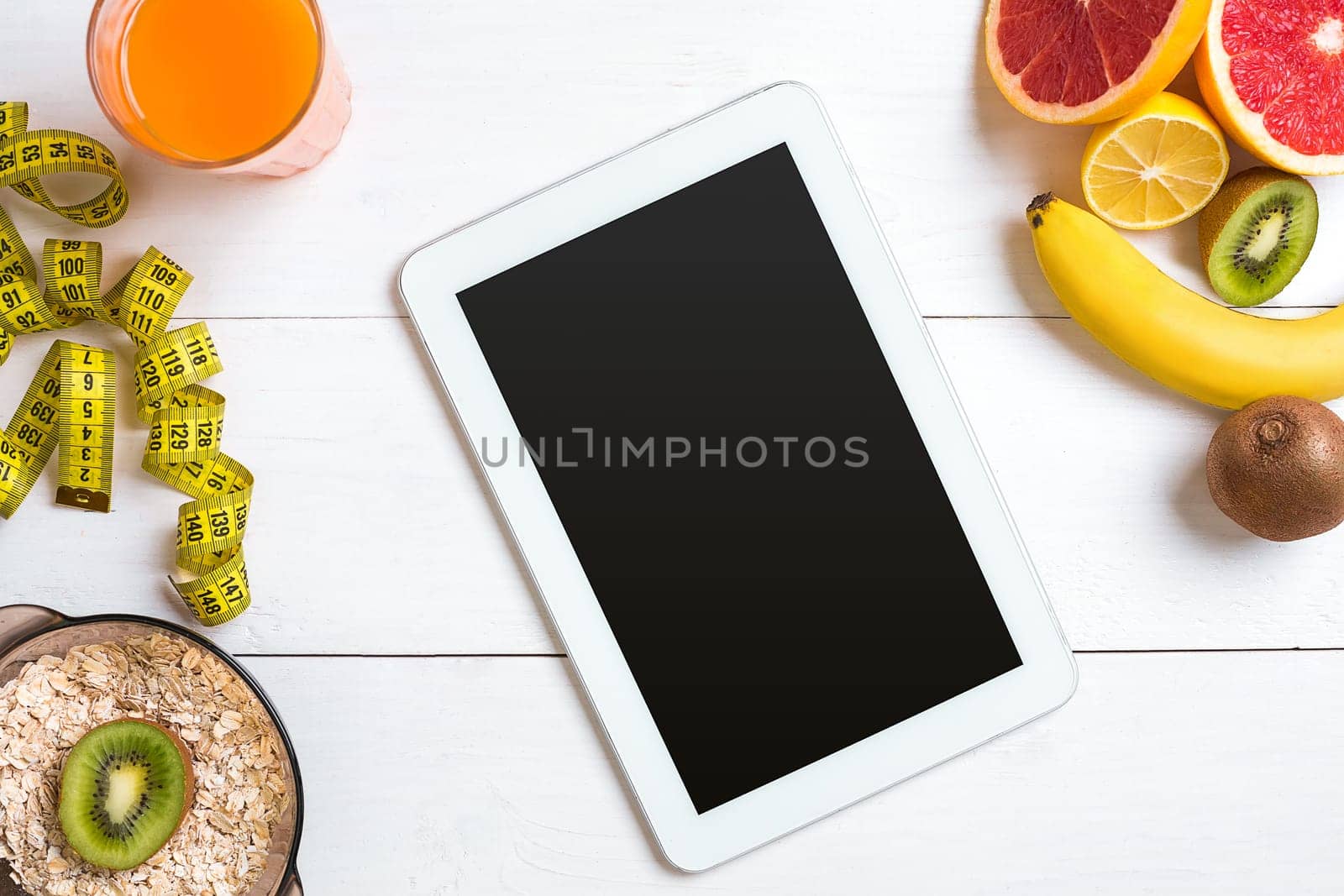 Fresh juice in glass from citrus fruits - lemon, grapefruit, orange, bowl of oatmeal on white wooden background, top view. White tablet with copy space