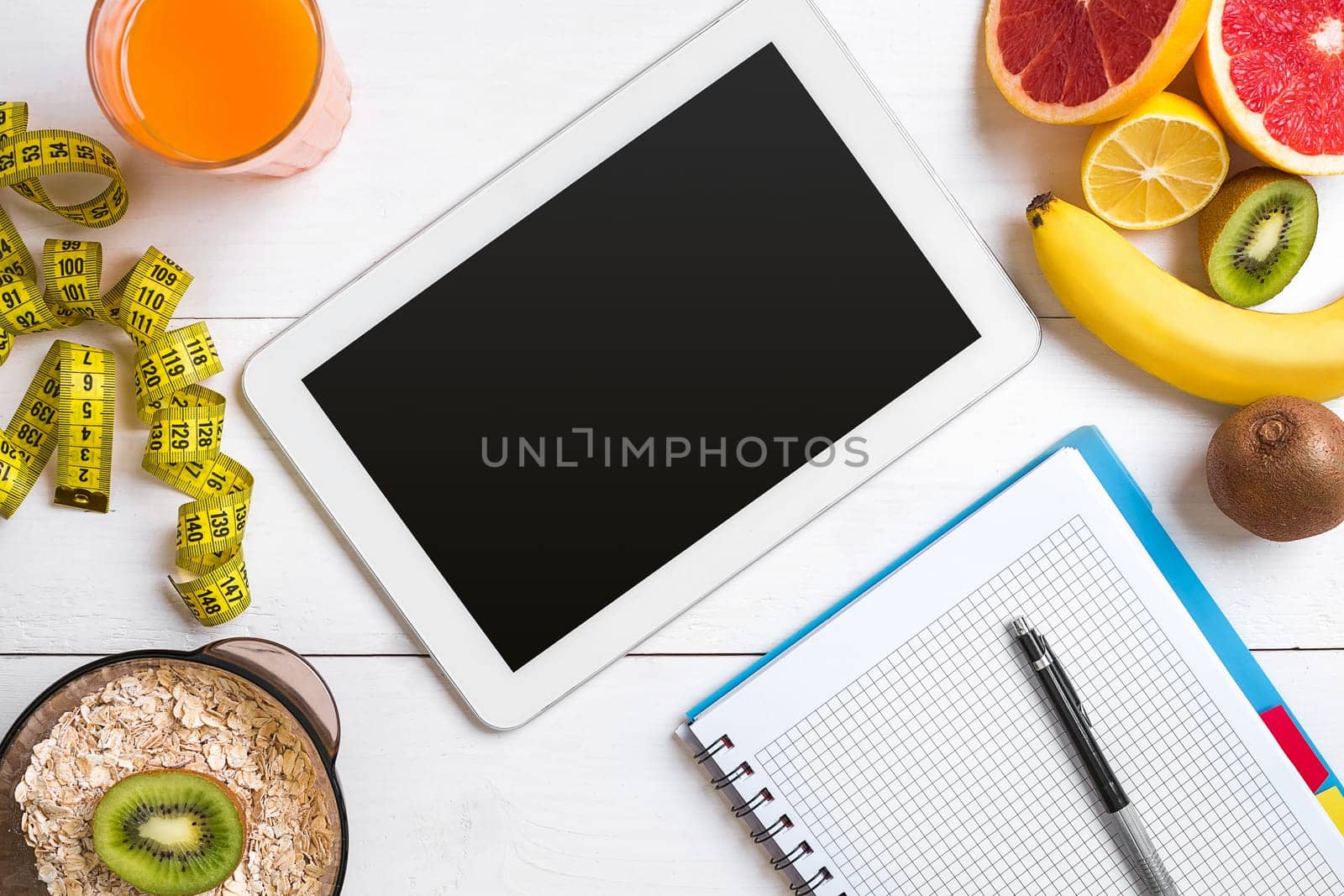 Fresh juice in glass from citrus fruits - lemon, grapefruit, orange, notebook with pencil on white wooden background, top view. White tablet with copy space