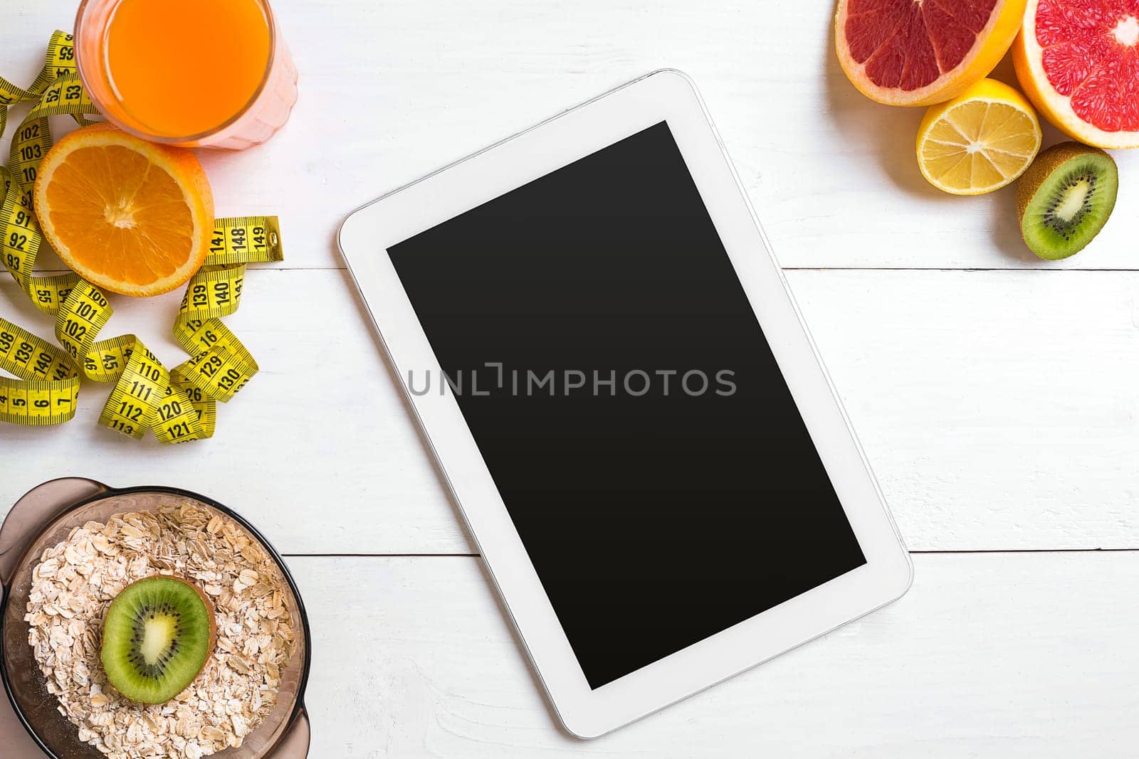 Fresh juice in glass from citrus fruits - lemon, grapefruit, orange, bowl of oatmeal on white wooden background, top view. White tablet with copy space