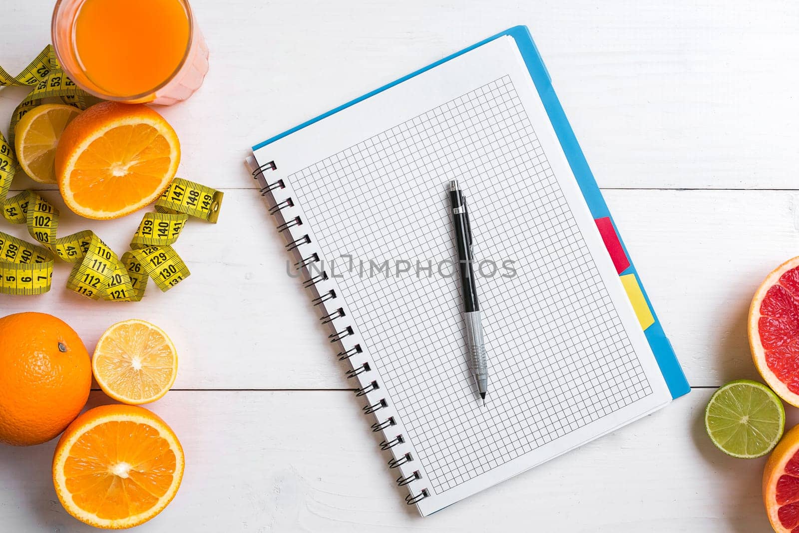 Fresh juice in glass from citrus fruits - lemon, grapefruit, orange, notebook with pencil on white wooden background, top view