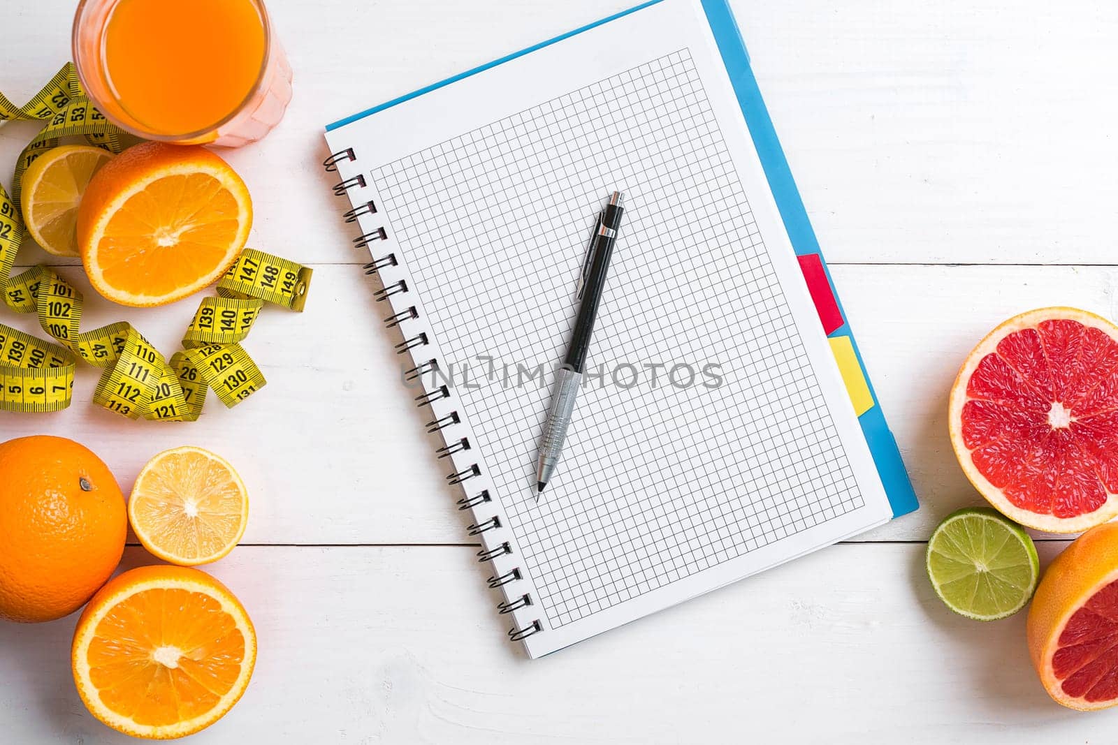 Fresh juice in glass from citrus fruits - lemon, grapefruit, orange, notebook with pencil on white wooden background, top view