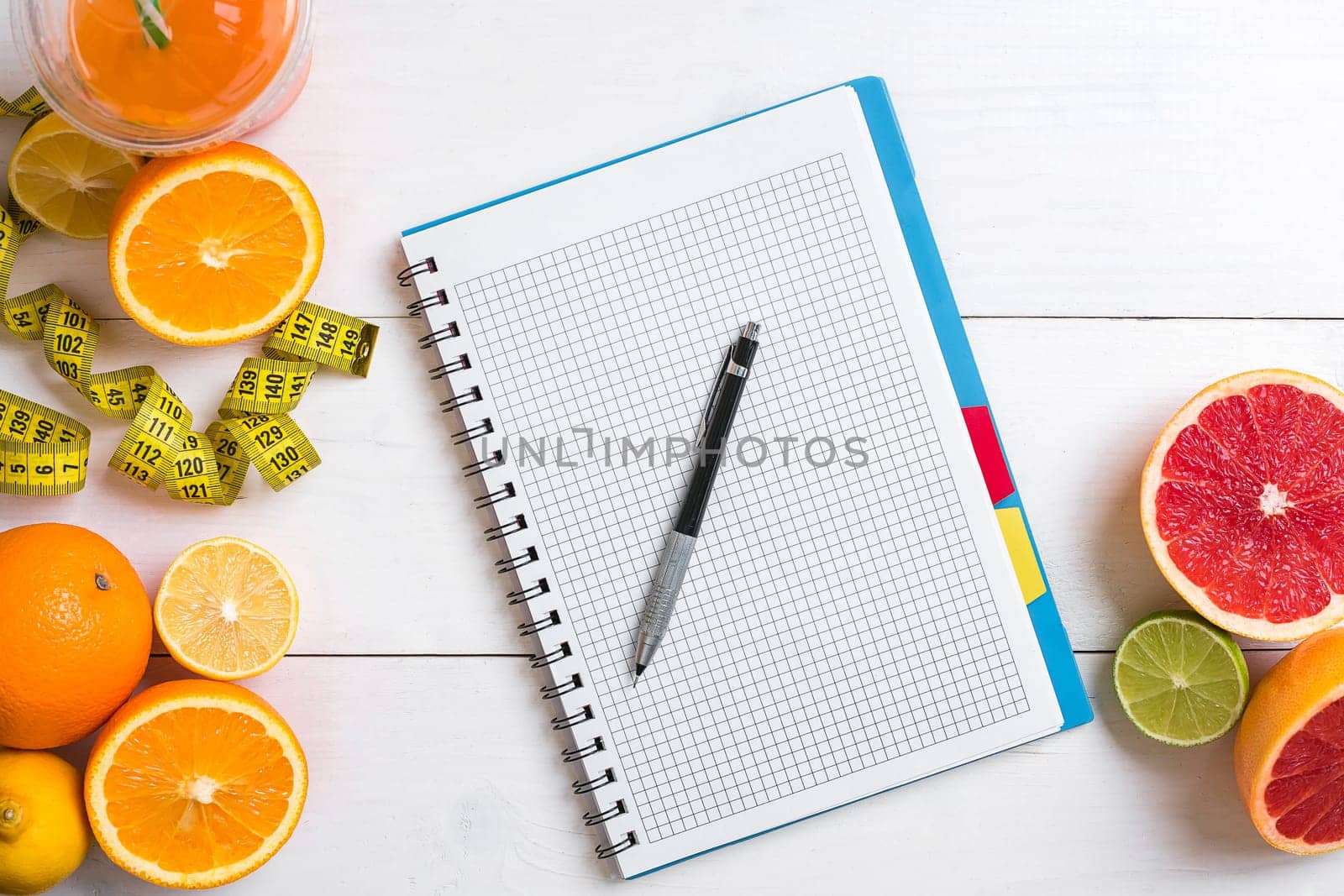 Fresh juice in glass from citrus fruits - lemon, grapefruit, orange, notebook with pencil on white wooden background, top view