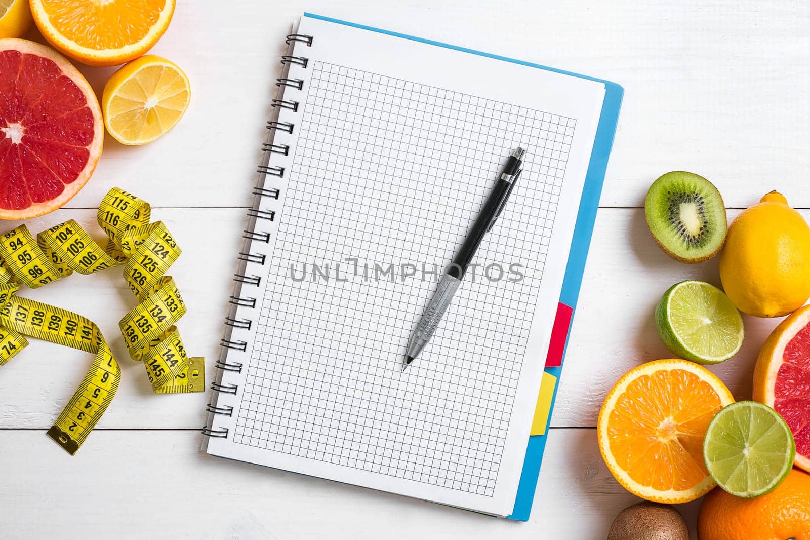 Fresh juice in glass from citrus fruits - lemon, grapefruit, orange, notebook with pencil on white wooden background, top view