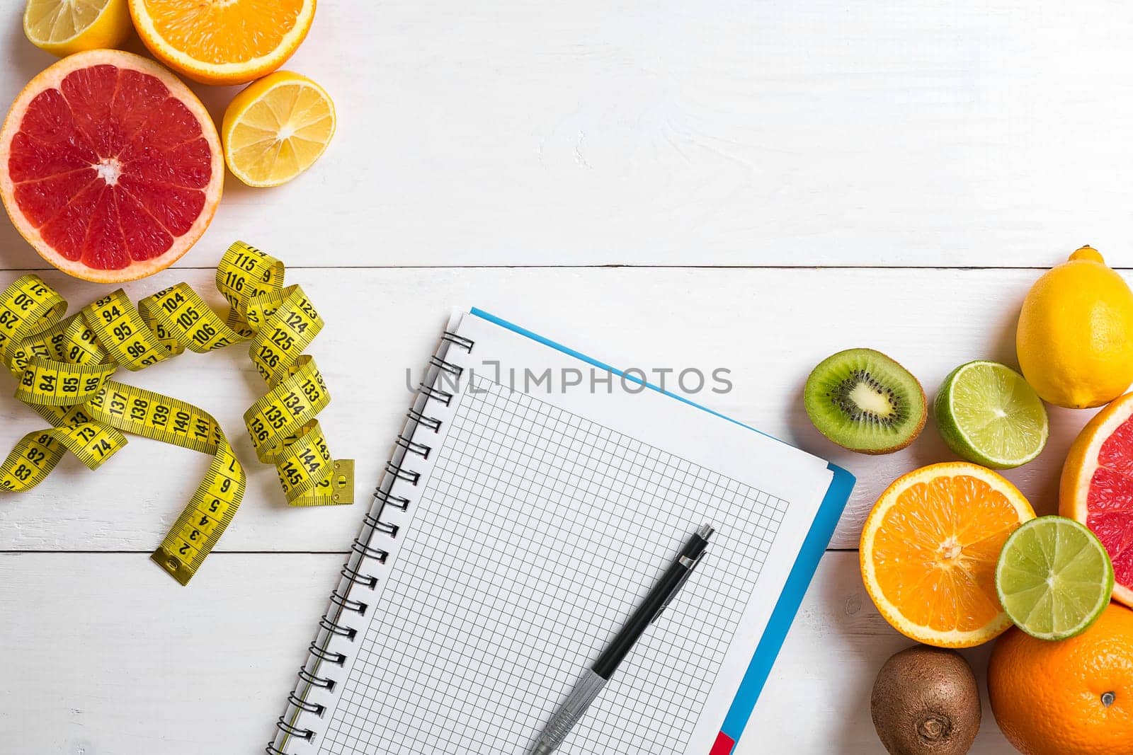 Fresh juice in glass from citrus fruits - lemon, grapefruit, orange, notebook with pencil on white wooden background, top view