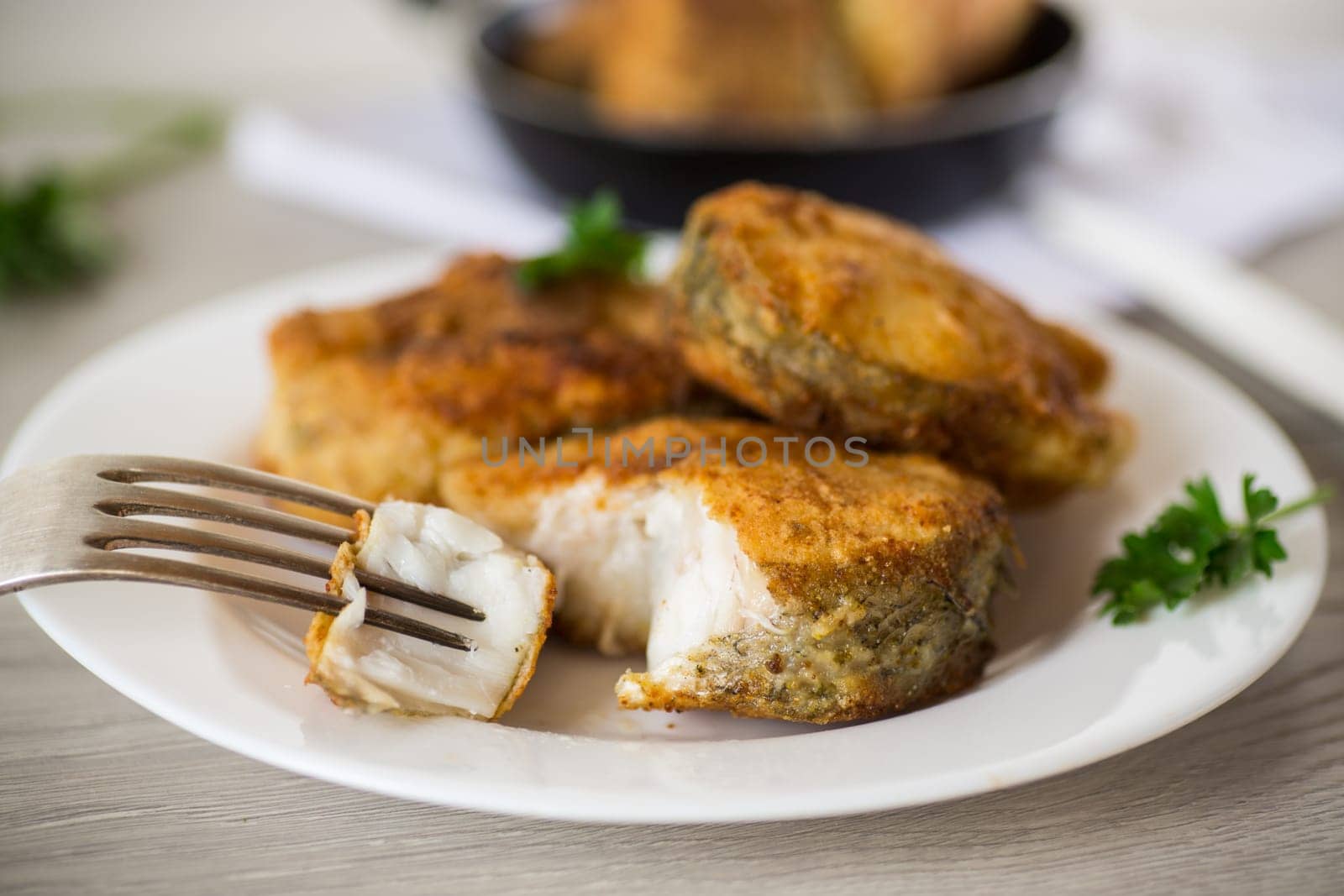 A piece of hake fish fried in a crispy crust, in a plate on a wooden table.