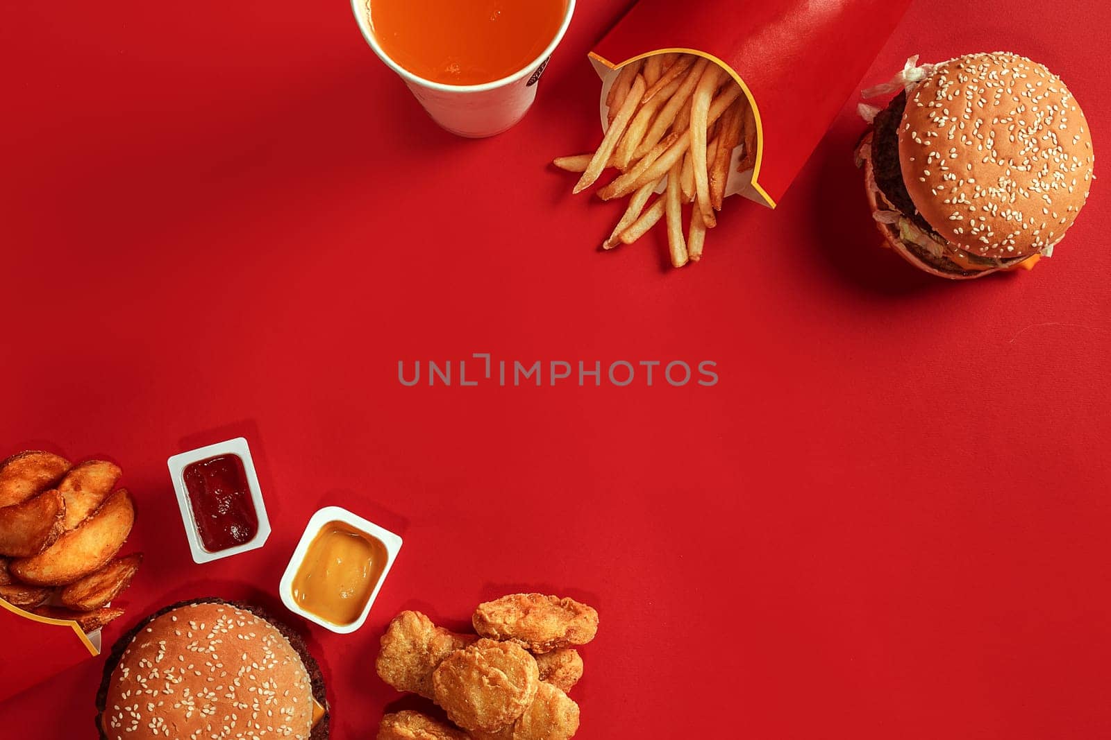 Fast food dish top view. Meat burger, potato chips and glass of drink on red background. Takeaway composition. Wrapped French fries, hamburger, mayonnaise and ketchup sauces on red desk. Copy space