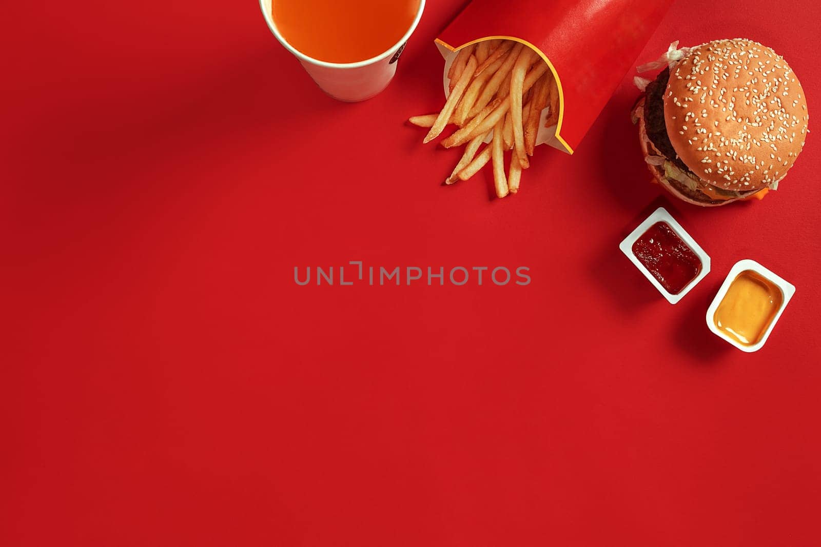 Fast food dish top view. Meat burger, potato chips and glass of drink on red background. Takeaway composition. Wrapped French fries, hamburger, mayonnaise and ketchup sauces on red desk. Copy space