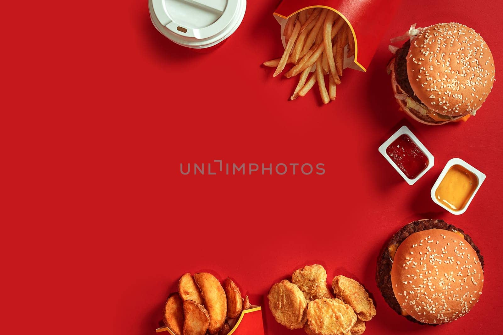 Fast food dish top view. Meat burger, potato chips and glass of drink on red background. Takeaway composition. Wrapped French fries, hamburger, mayonnaise and ketchup sauces on red desk. Copy space