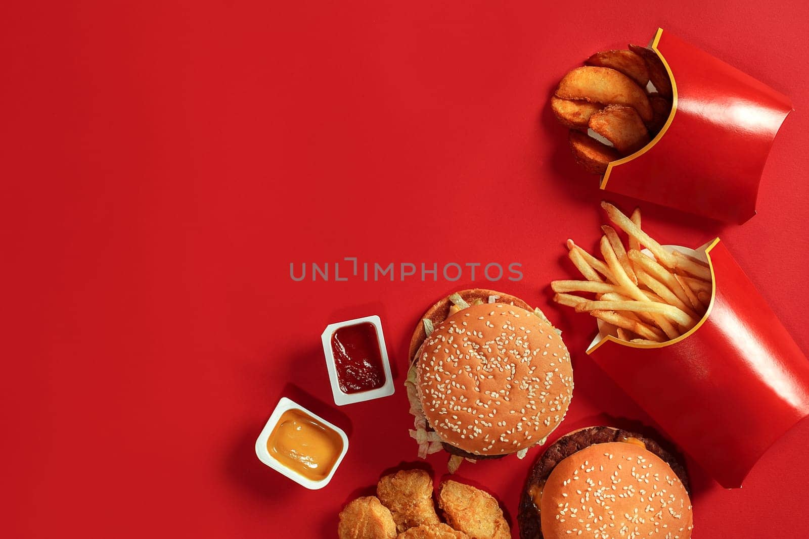 Fast food dish top view. Meat burger, potato chips and nuggets on red background. Takeaway composition. Wrapped French fries, hamburger, mayonnaise and ketchup sauces on red desk. Copy space