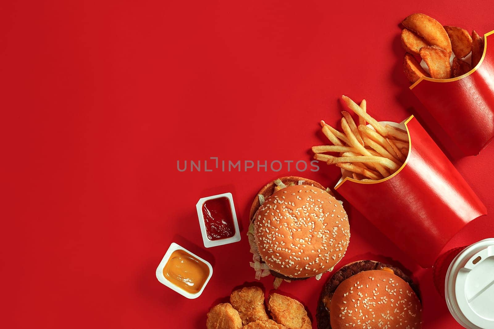 Fast food dish top view. Meat burger, potato chips and glass of drink on red background. Takeaway composition. Wrapped French fries, hamburger, mayonnaise and ketchup sauces on red desk. Copy space