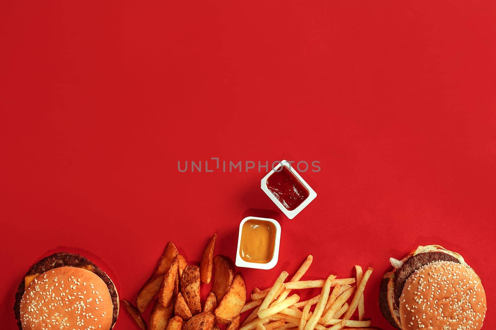 Fast food dish top view. Meat burger, potato chips and nuggets on red background. Takeaway composition. Wrapped French fries, hamburger, mayonnaise and ketchup sauces on red desk. Top view. Copy space