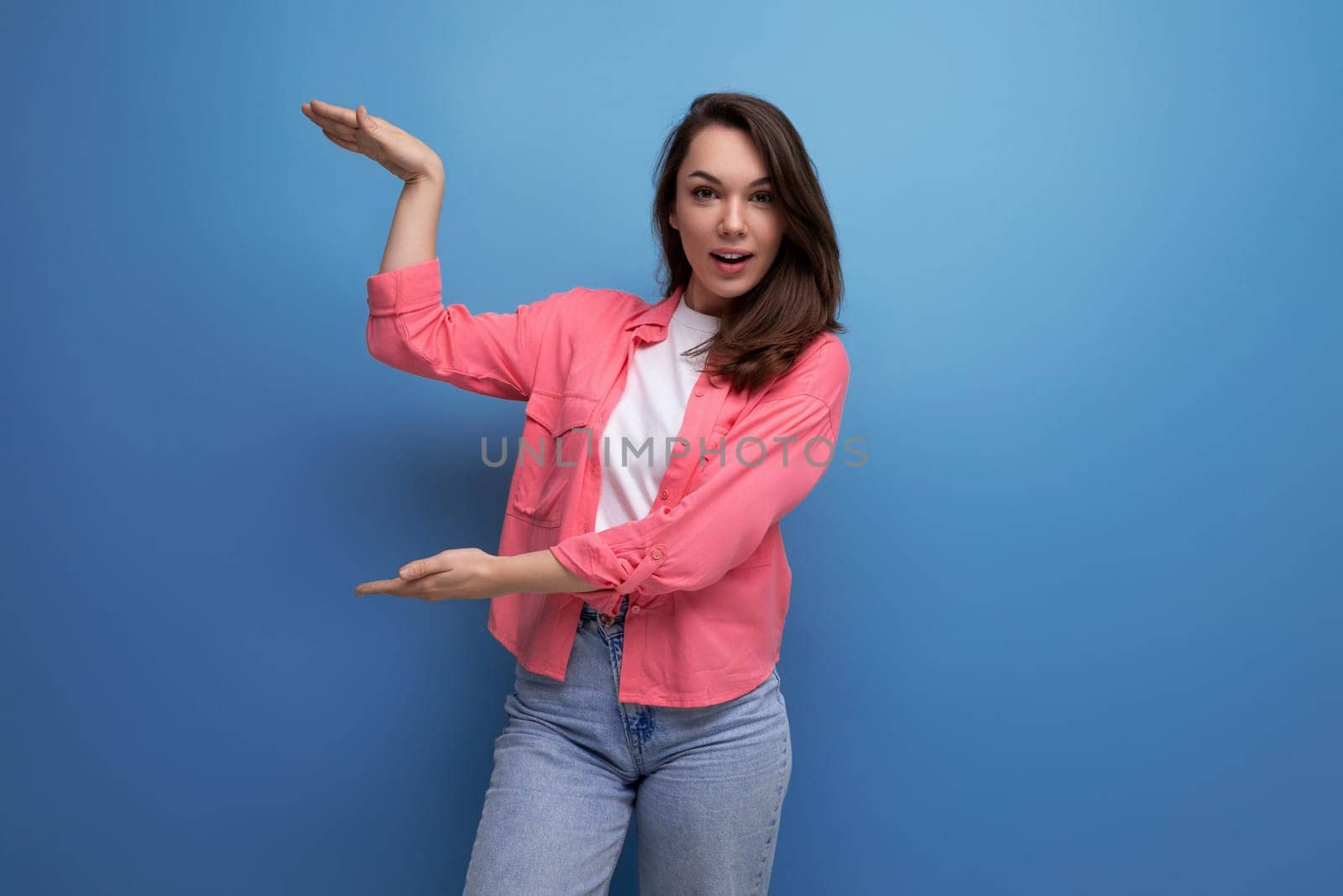 portrait of successful european young female adult with shoulder length hair in pink shirt showing joy with hands on studio background with copy space.