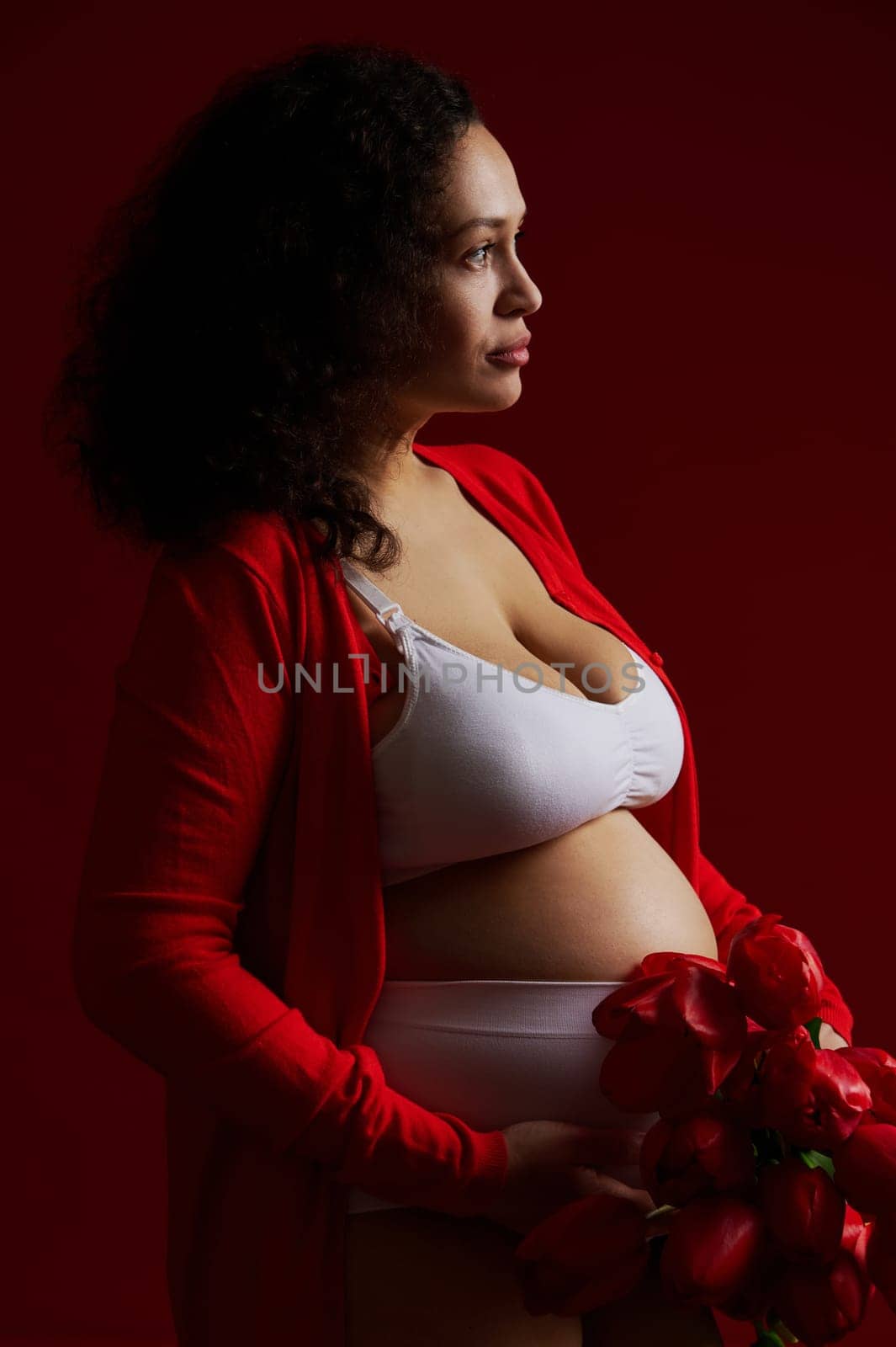 Beautiful multi ethnic pregnant woman holding a bunch of red tulips, posing bare belly over dark red background by artgf