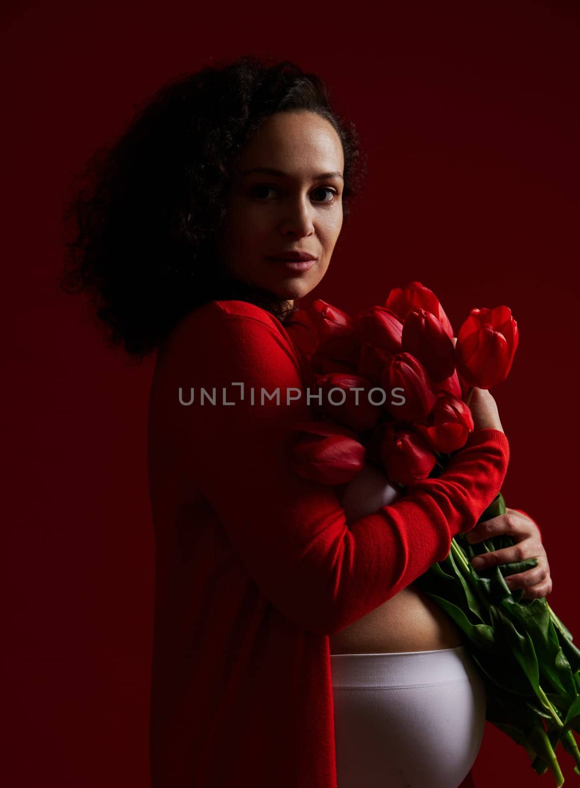 Authentic adult pregnant woman holding a bouquet of tulips, looking confidently at camera, isolated on dark background by artgf