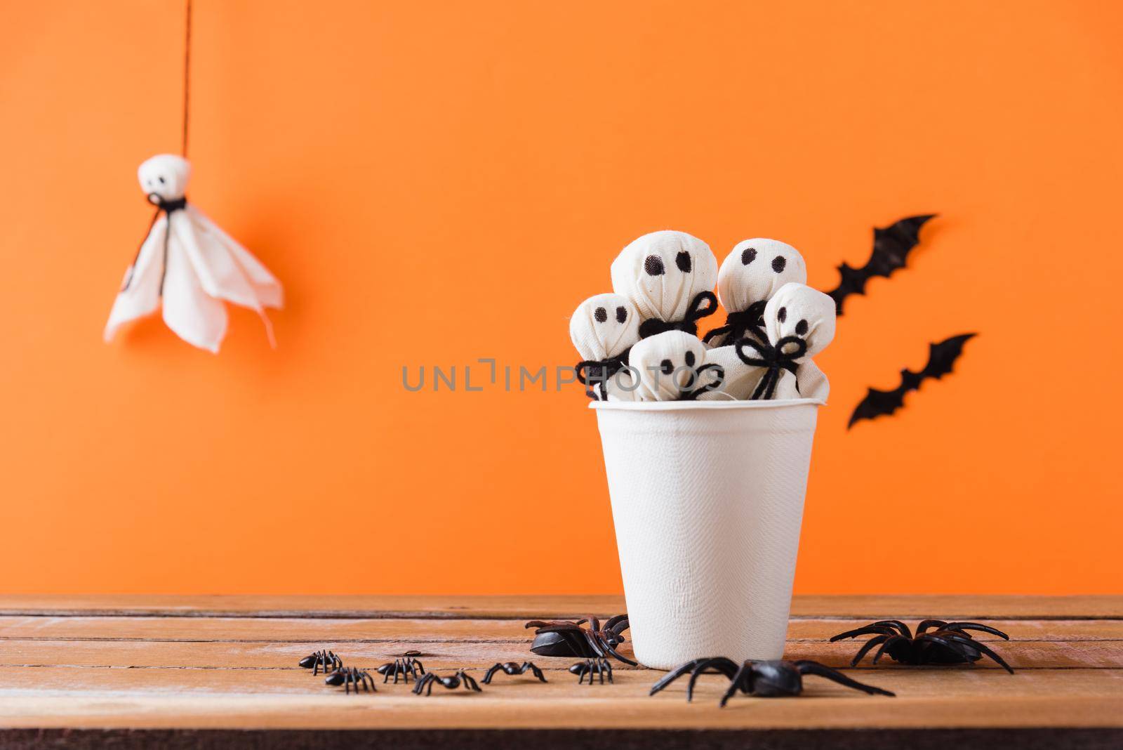 Funny Halloween day decoration party, White ghost crafts scary face head in paper cup on wood table, studio shot isolated on orange background have spider and bats, Happy holiday DIY concept