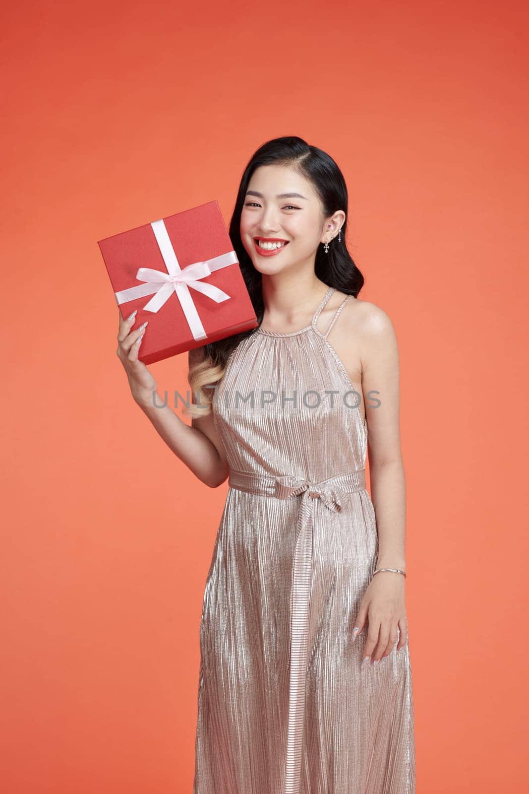 Elegant young woman in glamorous dress holding wrapped gift box isolated on red by makidotvn
