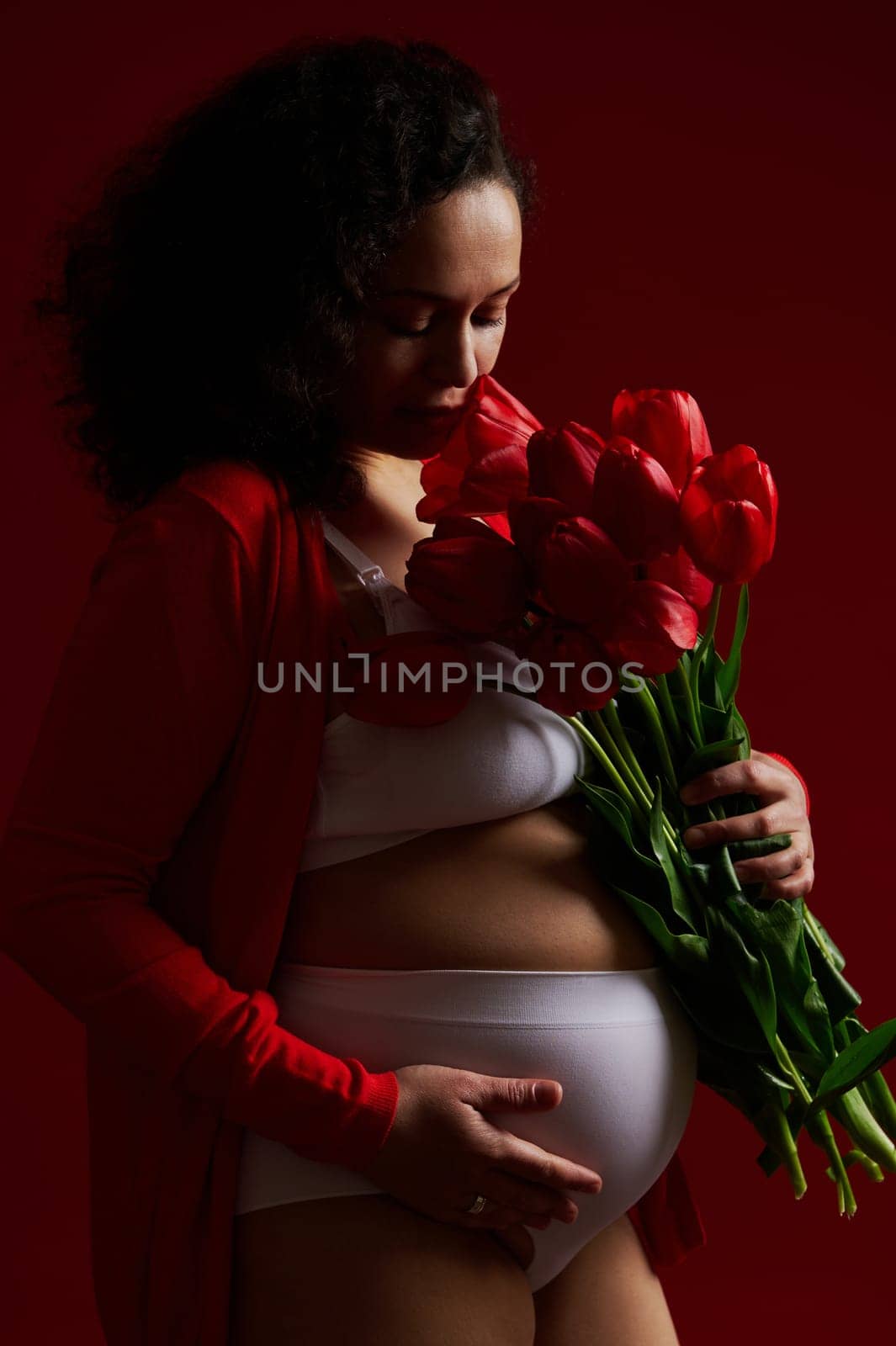 Pregnant woman, expectant mother sniffing a bouquet of red tulips, gently stroking her belly, on isolated background by artgf
