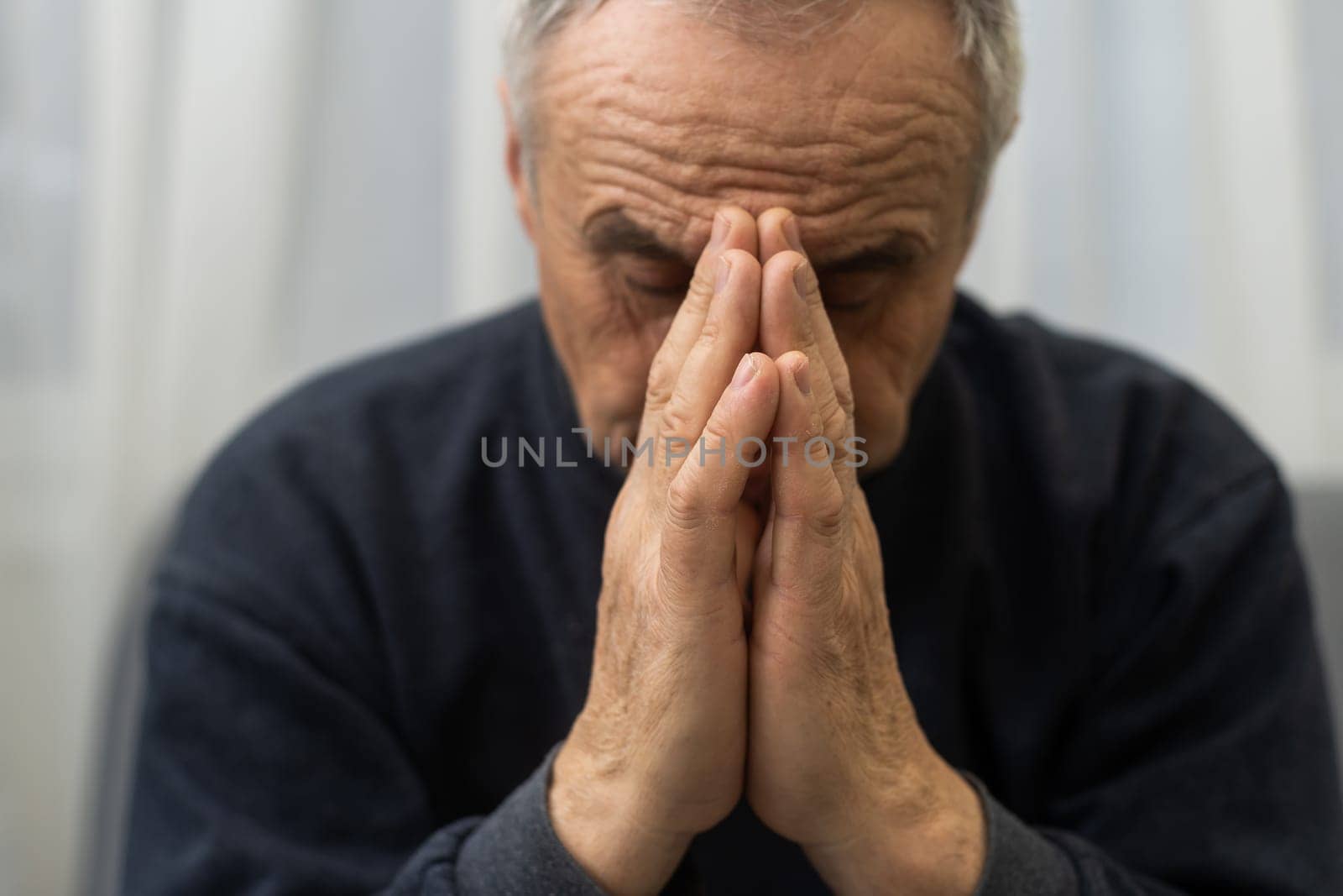 Please, Lord. Nervous worried aged Caucasian man praying on couch at home. Stressed white European senior citizen sitting on sofa, begging for forgiveness or asking God for help in difficult situation