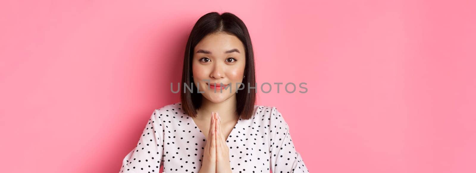 Beauty and lifestyle concept. Close-up of cute asian woman smiling, showing thank you gesture, holding hands in pray, standing over pink background.