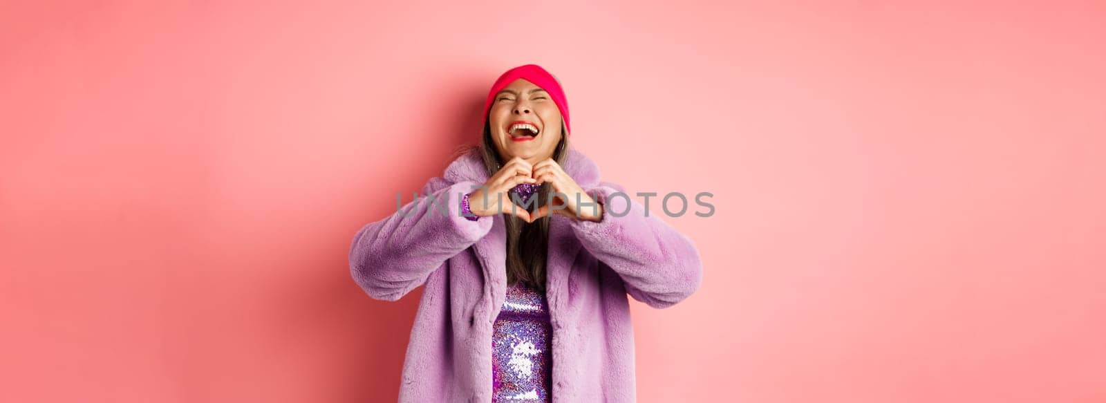 Valentines day and shopping concept. Happy asian woman in stylish outfit showing heart sign and laughing carefree, standing in faux fur against pink background.