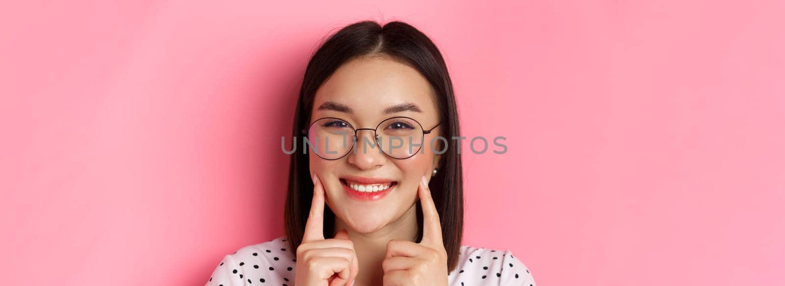 Beauty concept. Headshot of adorable asian girl in trendy glasses smiling, poking cheeks and showing cute dimples, standing over pink background.