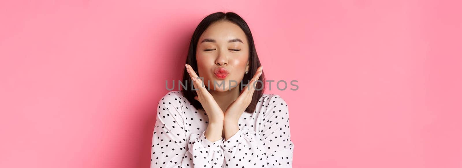 Beauty and skin care concept. Close-up of beautiful asian girl close eyes, pucker lips for kisses, showing cute perfect face, standing over pink background by Benzoix