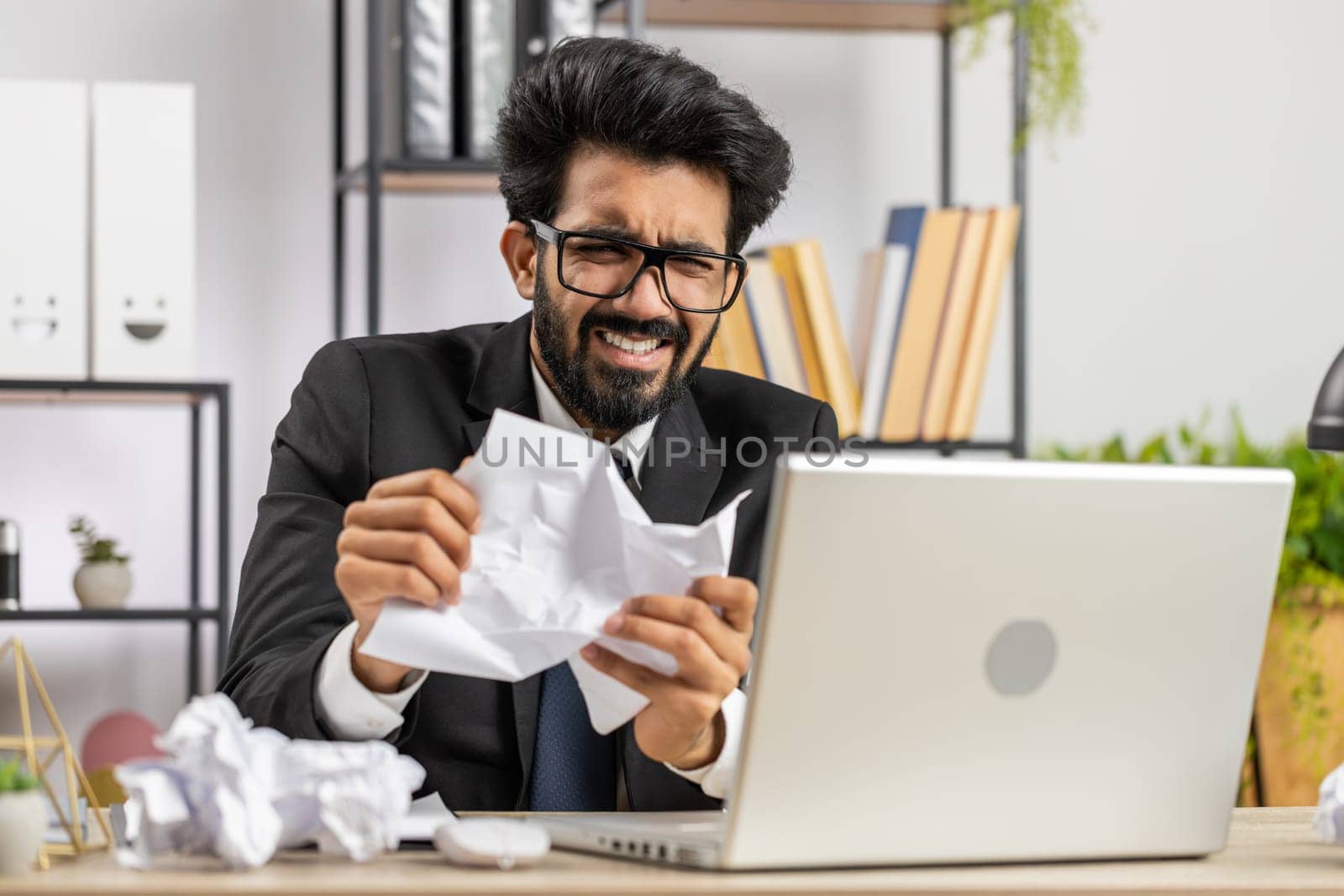 Angry furious indian man working at office throwing crumpled paper, having nervous breakdown at work, migaine, headache, stress management, mental distress problems, losing temper, reaction on failure