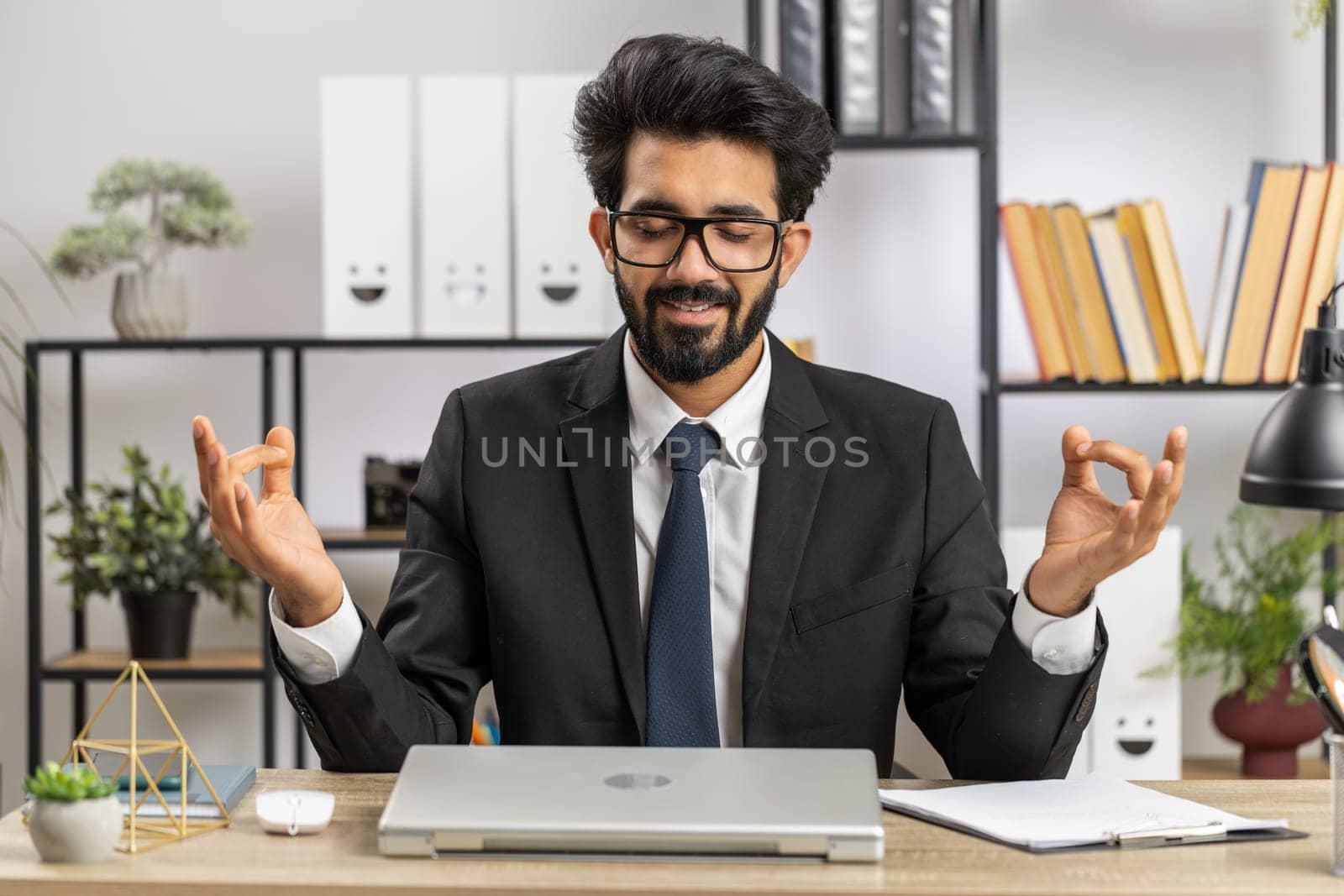 Indian businessman working on laptop computer, meditating, doing yoga breathing exercise in lotus position at home office. Calm serene freelancer arab man taking break. Busy occupation. Peace of mind
