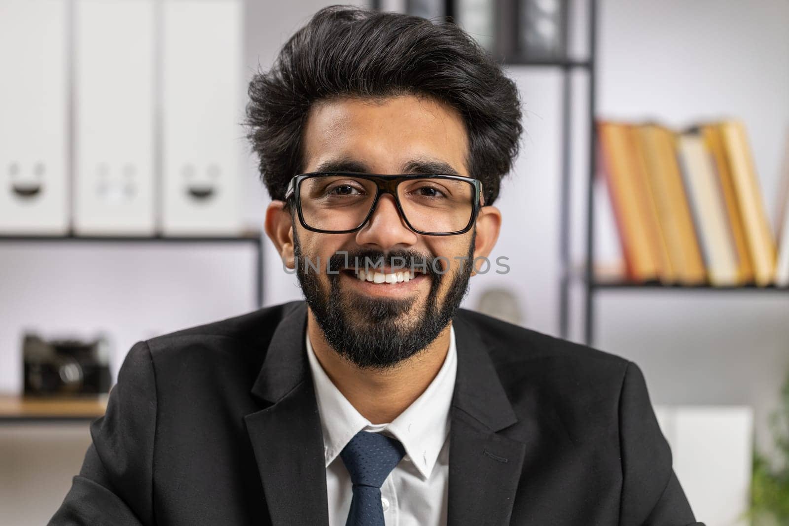 Portrait of happy smiling bearded indian businessman at modern home office workplace desk looking at camera. Male hindu in formal suit. Handsome man Remote distant online working on laptop
