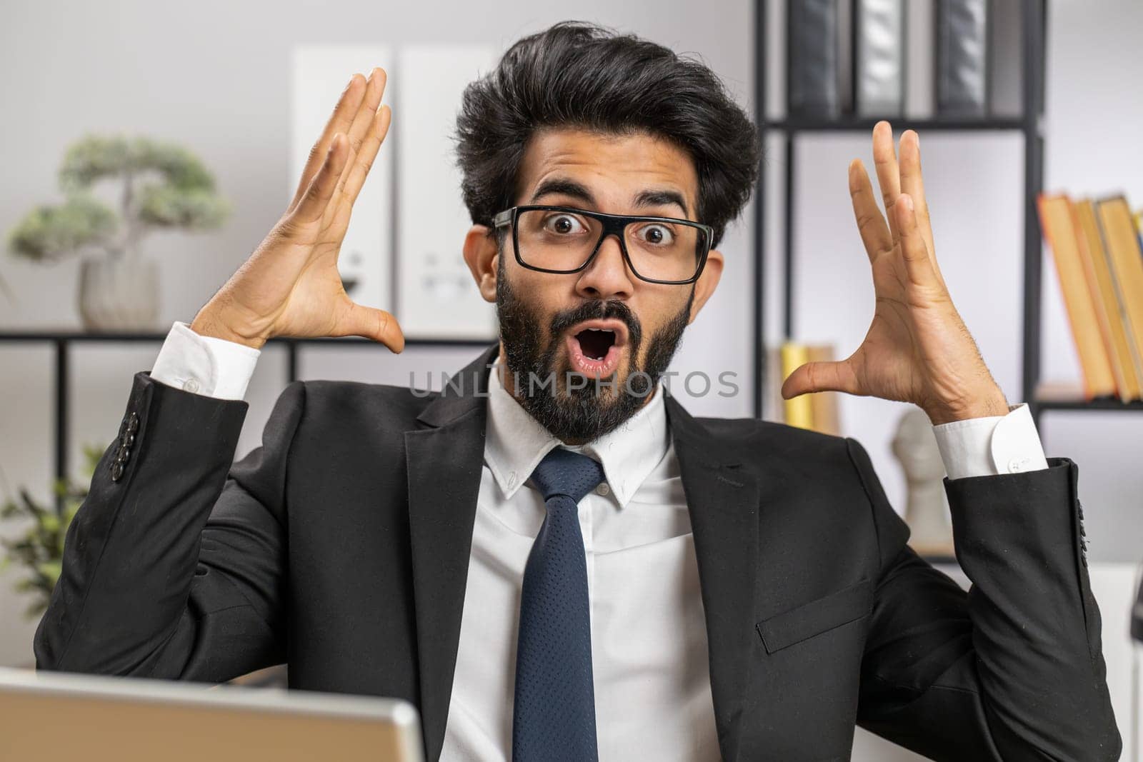 Oh my God, Wow. Indian businessman working at home office workplace, looking at camera shocked by sudden victory, game winning, lottery goal achievemen, good news. Professional manager freelancer man