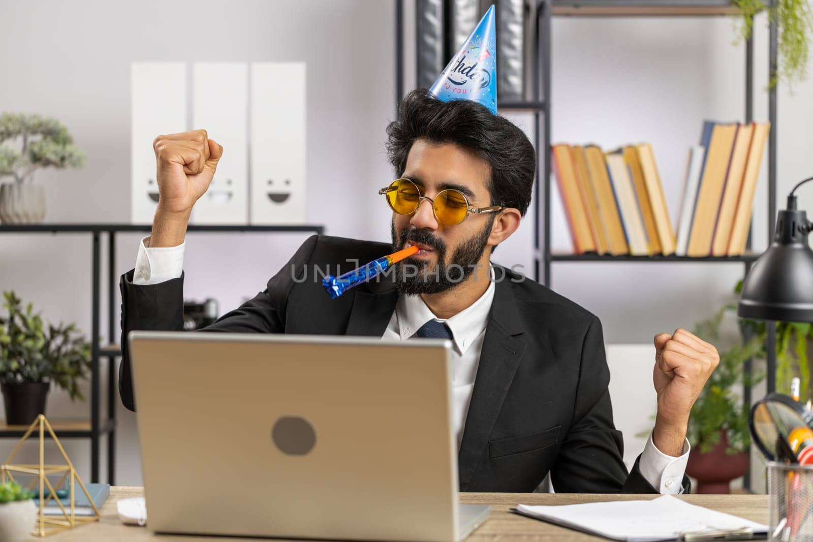 Excited lucky business man in sunglasses, festive cap hat cone blow pipe at workplace dancing victory dance, weekend. Rejoicing developing project. Celebrating birthday. Lottery jackpot. Office people