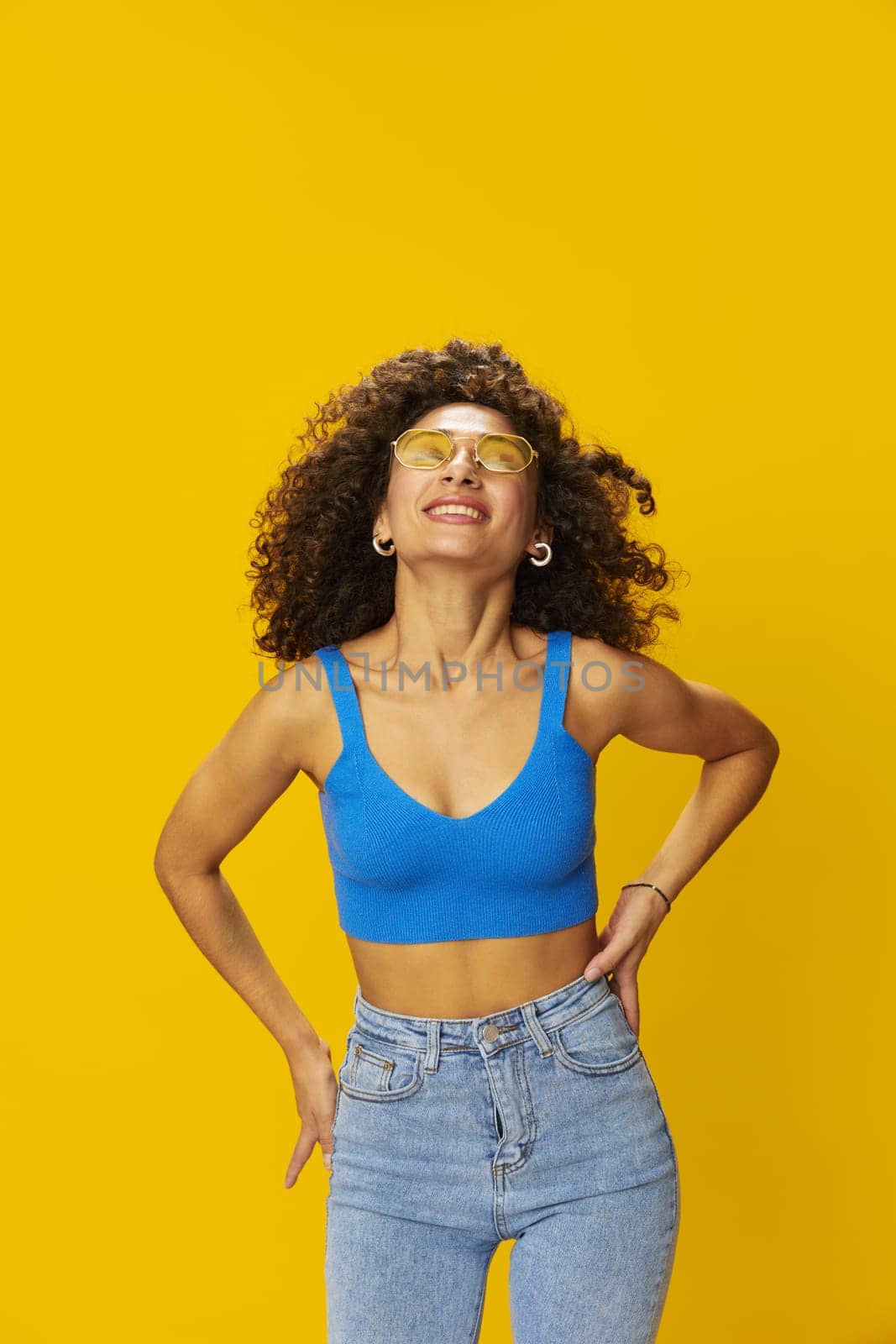 Woman with curly afro hair in a blue T-shirt on a yellow background dancing flying hair with sunglasses yellow, hand signs, look into the camera, smile with teeth and happiness, copy space. High quality photo