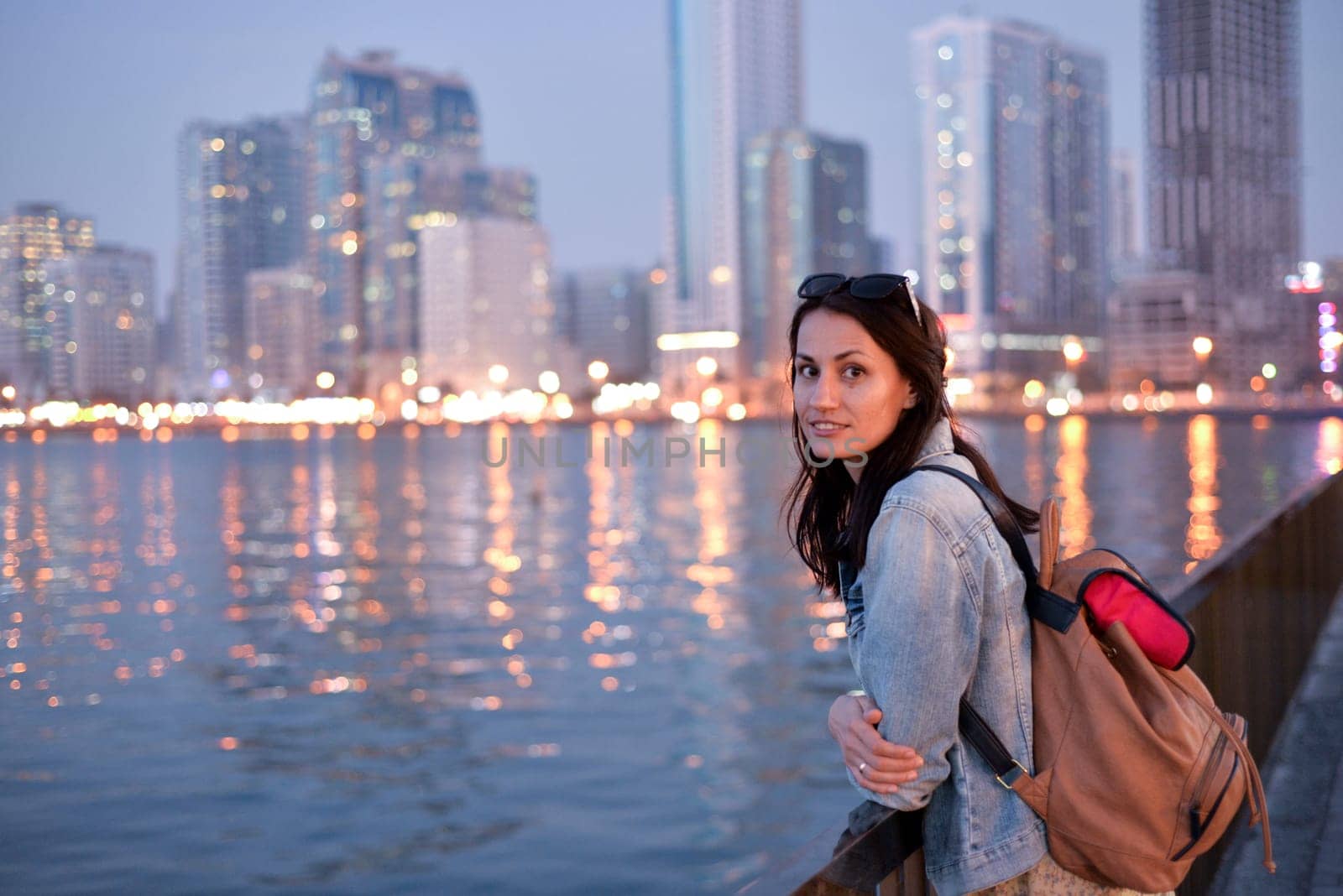 A tourist girl with a backpack on her shoulders enjoys a view of the modern skyscrapers of the Sharjah marina at night.