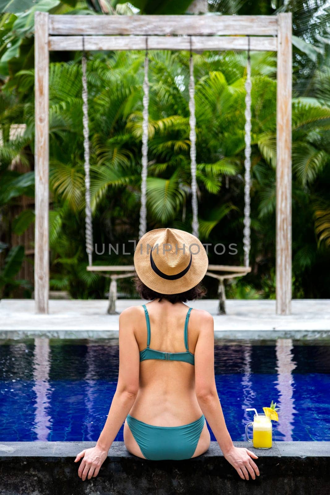 Rear view of woman in bikini sitting on swimming pool ledge relaxing looking at tropical view during vacation. Vertical by Hoverstock