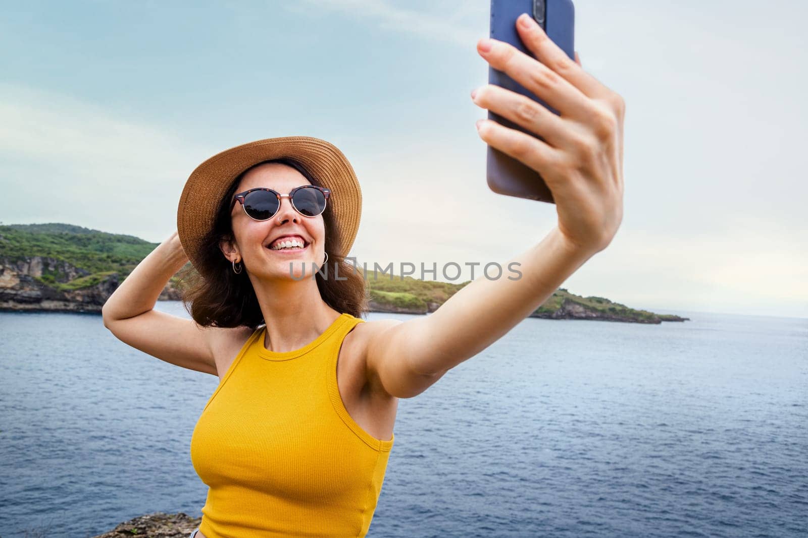 Young female tourist traveler taking selfie with phone in seascape during summer vacation in Nusa Penida, indonesia. Travel, social media and technology concepts.