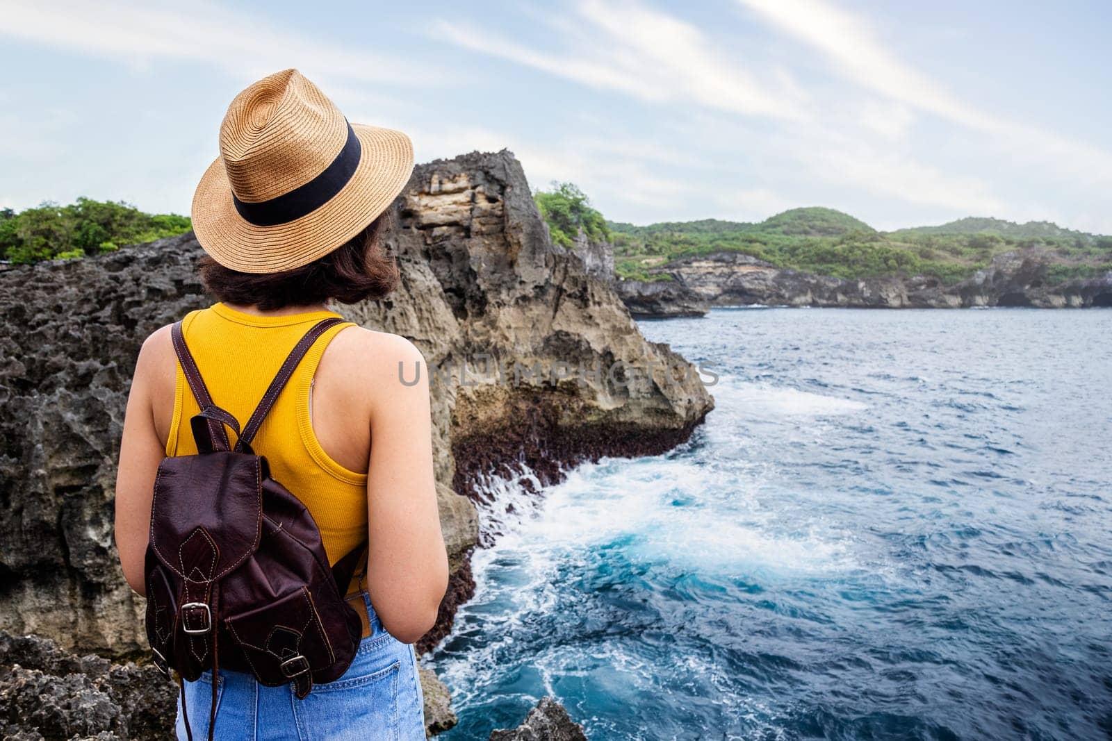 Rear view of young woman with hat and backpack contemplating nature. Female looking at ocean view during summer vacation. Angels Billabong ,Nusa Penida island, Bali, Indonesia. Traveling, freedom and wellness concept.